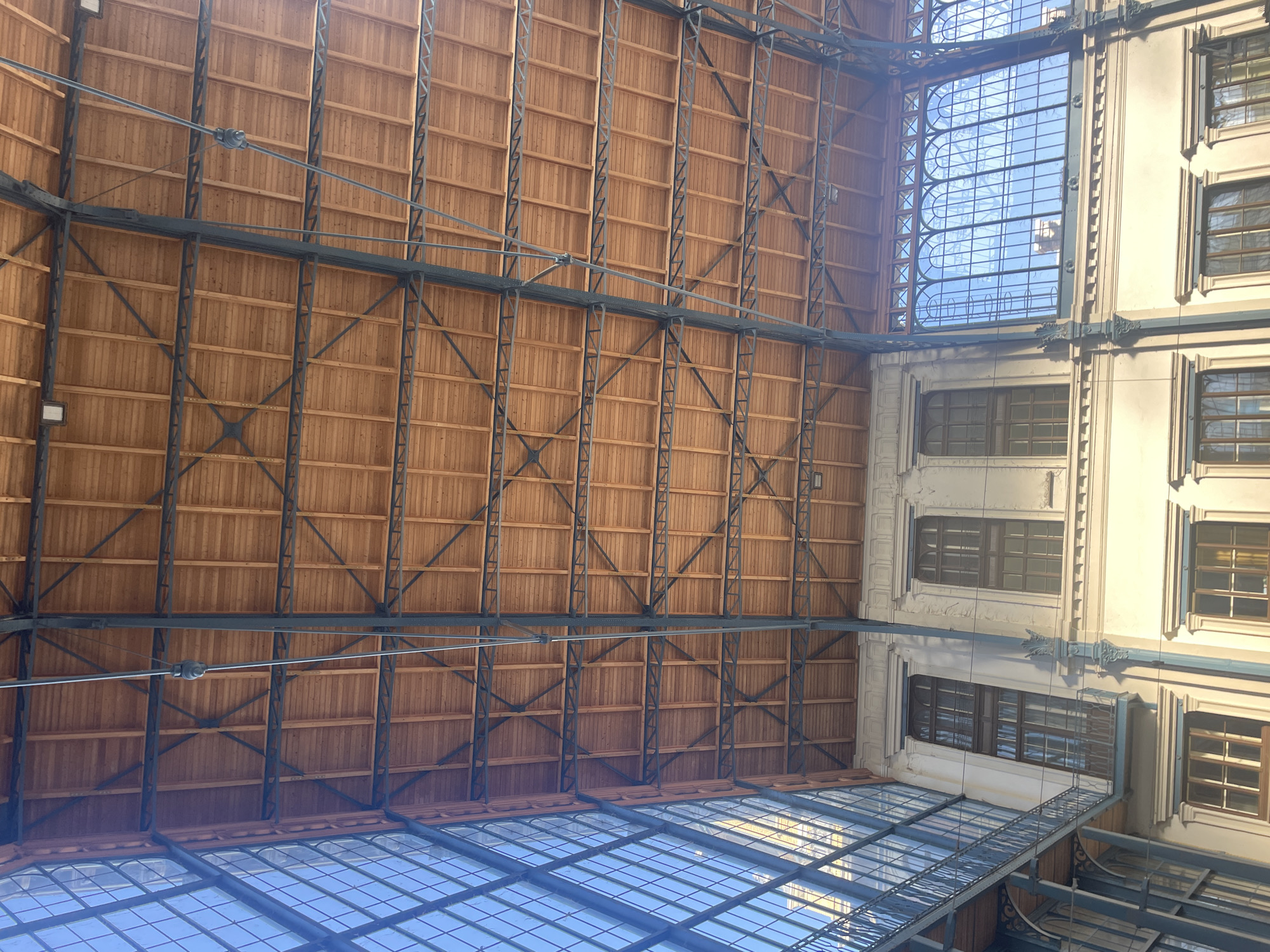 This image depicts the interior of a building in Budapest, Hungary, specifically the Párisi Udvar. The view is directed upwards towards the ceiling and adjacent building structure. The ceiling is constructed from light brown wooden planks arranged in a grid pattern, reinforced by dark metal beams in a truss design. A glass-paneled roof is visible below the wooden structure, reflecting light from the sky. To the right, the exterior facade of an adjacent building is seen, featuring white or light-colored walls with multiple window openings, exhibiting classical architectural details and embellishments. The windows have dark frames, and the building's facade appears to be ornamented with decorative molding.