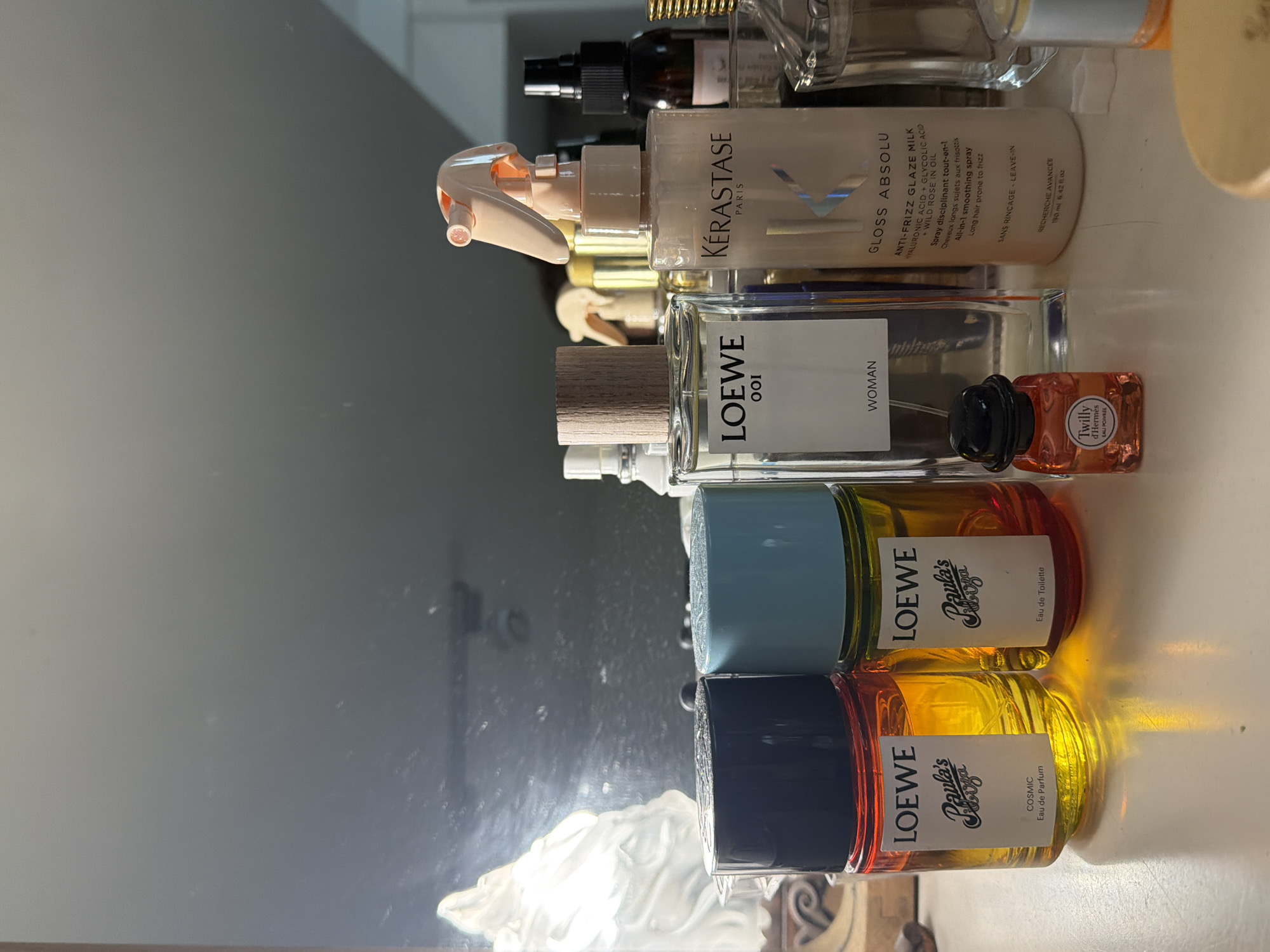 The image shows a collection of beauty products stacked vertically on a white surface. At the top is a Kérastase Gloss Absolu Anti-Frizz Glaze Milk spray. Below it is a bottle of LOEWE 001 Woman perfume, followed by a miniature Twilly d'Hermès eau poivrée fragrance. Underneath those are two bottles of LOEWE Paula's Ibiza perfume, one Eau de Toilette with a blue cap, and one Eau de Parfum Cosmic with a black cap. The items are stacked in front of a mirror. The lighting is bright, casting strong shadows. The location in the city of Buenos Aires, Argentina, is not discernible from this image.