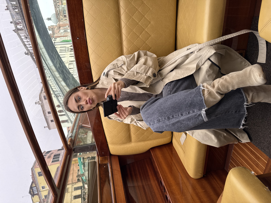 A light-skinned female is seated inside a polished wooden water taxi or private boat on a canal in Venice, Italy. She is depicted from the waist up, looking directly at the camera with wide eyes and pursed lips. She wears a beige trench coat over a brown V-neck sweater, dark grey wide-leg jeans with frayed hems, and light beige boots. Both hands hold a small, black electronic device with a flip-out screen, angled as if in use for recording or photography. The boat's interior features yellow diamond-quilted upholstered seating, polished wooden panels, and a dark grey carpeted floor. Through the clear windows, an overcast Venetian cityscape is visible, including multi-story traditional buildings lining the canal, a grey stone arched bridge with a balustrade spanning the waterway, and a large domed architectural structure in the distant background.