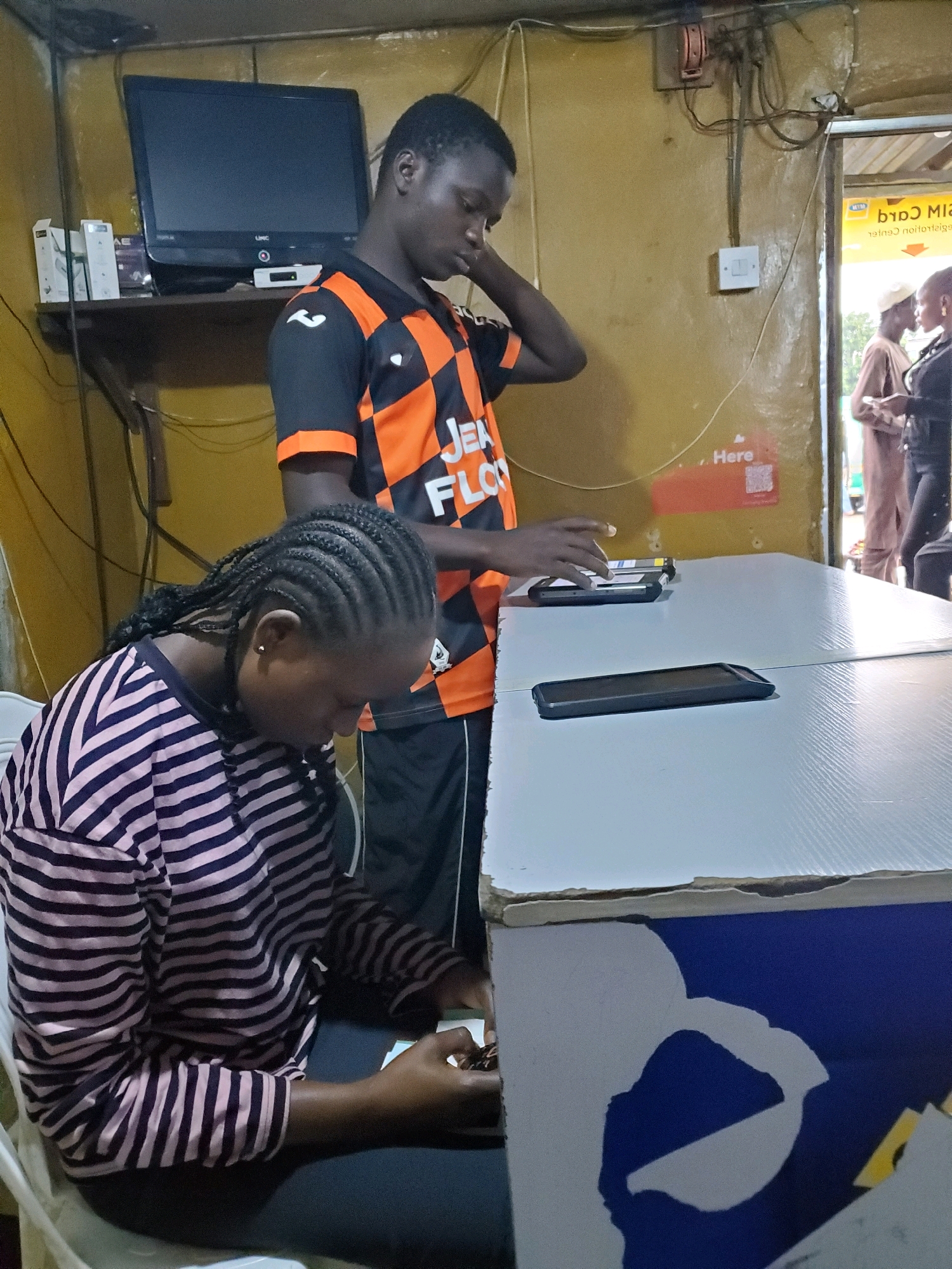 In Gura Top, Nigeria, two individuals are interacting at a counter inside a small, indoor establishment. A young man, wearing a black and orange checkered shirt with "JEA FLO" written on it, stands on the right, with his right hand touching the back of his neck. In front of him, a young woman with braided hair is seated, looking down at a mobile phone in her hands. She is wearing a pink and navy striped long-sleeved shirt. On the counter in front of them are two mobile phones, one black and one silver, and a tablet. The counter surface is white and textured, and the side of the counter has a blue and white graphic. In the background, a black television is mounted on a shelf above some white boxes. Electrical wires and a light switch are visible on the yellow-painted wall. To the right, an open doorway shows two other individuals in the distance, one wearing a brown garment and a cap, and another in a black outfit. A yellow sign with black text is partially visible above the doorway.