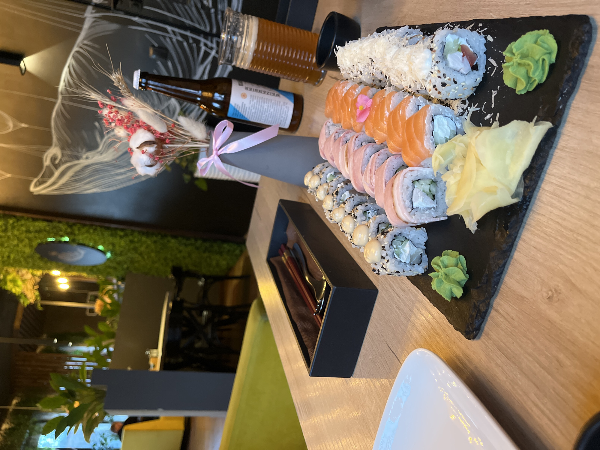 This close-up, high-angle shot captures a meticulously arranged meal on a light wooden table, likely in a restaurant or cafe in Izhevsk, Russia. The central focus is a large black slate board showcasing an impressive assortment of sushi rolls. Several varieties are present, including rolls topped with vibrant orange salmon slices, others adorned with a white shredded topping (possibly grated cheese or coconut), and various rolls featuring sesame seeds or small white beans/peas. Neatly accompanying the sushi are a dollop of bright green wasabi and a pile of pale yellow pickled ginger.
To the left of the sushi platter, a brown glass bottle of beer is visible, with the word "WEIZENBIER" clearly printed on its label. A glass containing a golden-brown beverage, likely beer, stands nearby. A small, decorative bouquet adds a touch of elegance, featuring dried pink flowers, fluffy white elements (possibly cotton), and wheat stalks, tied with a pink ribbon and placed in a dark gray vase.
Further down the table, a black rectangular box holds chopsticks, ready for use. A white, triangular-shaped plate is partially visible in the foreground. The background is softly blurred, revealing a dark wall decorated with abstract white line drawings and a vibrant green wall that appears to be covered in artificial plants or moss, suggesting an indoor, possibly evening, setting. No people are visible in the frame, but the setup implies a relaxing dining experience.