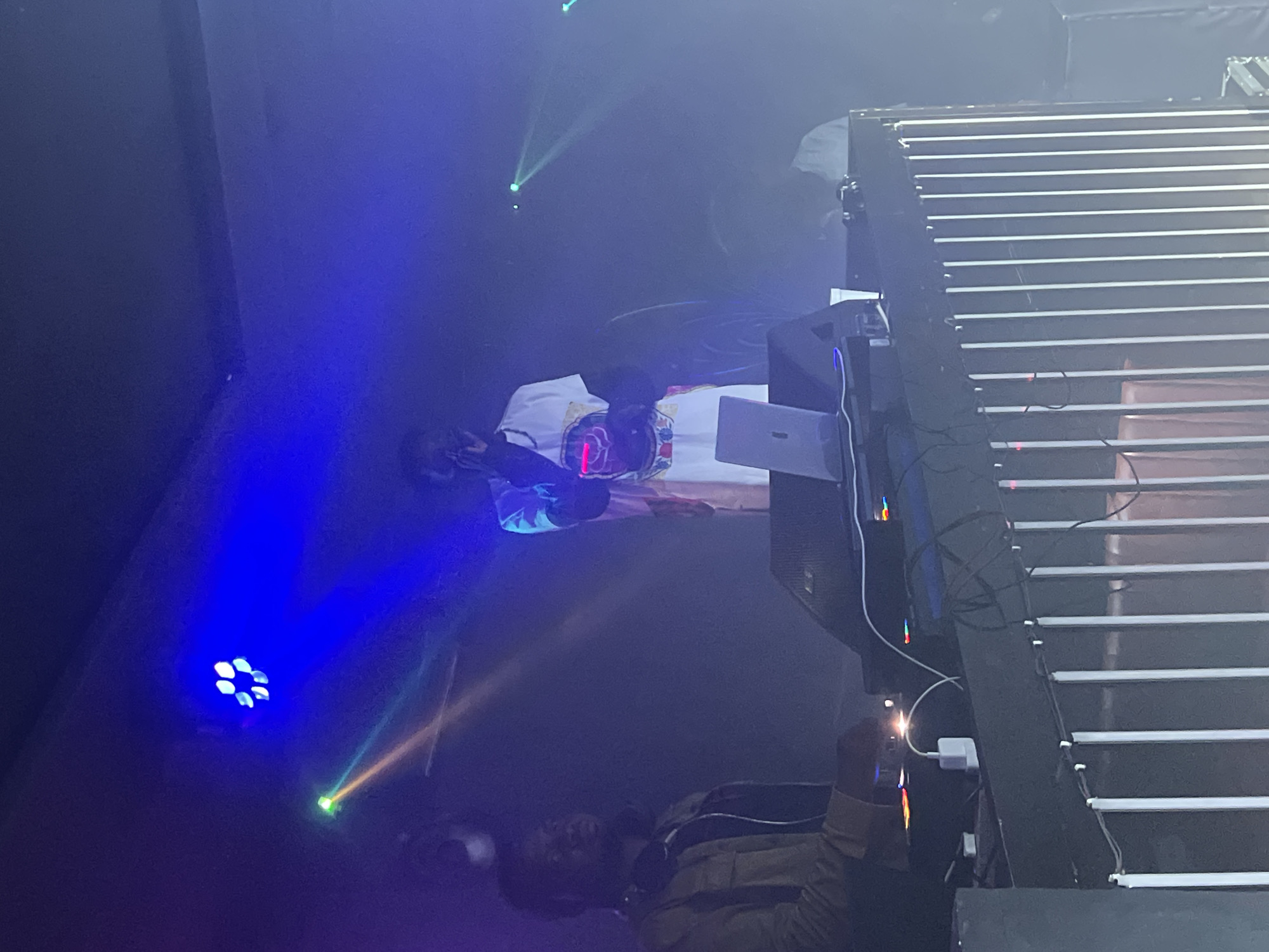 A concert stage is shown, illuminated by blue and green stage lights. A DJ with a white shirt is standing behind the DJ table. Another man is standing in the right corner wearing a brown jacket and facing away from the camera. Wires are hanging down from the scaffolding. The location is unknown.