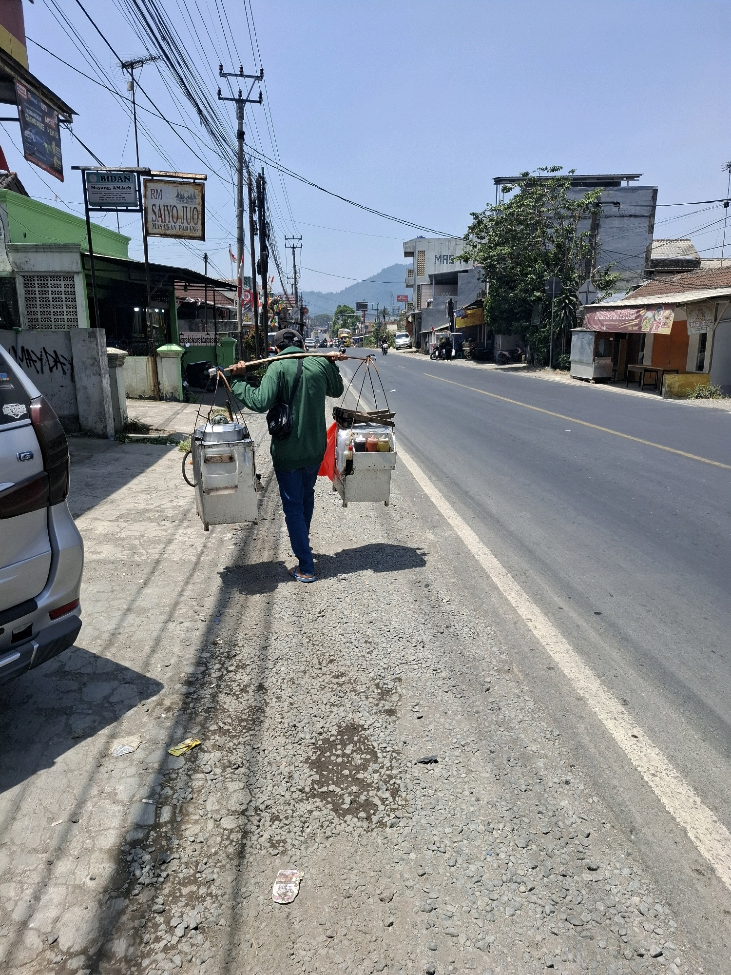 A street vendor walks along the side of a paved road in Selaawi, Indonesia, carrying a yoke across his shoulders. From the yoke hang two insulated containers, one on each side, likely containing food or drinks for sale. The vendor wears a green long-sleeved shirt, dark blue jeans, and flip-flops. His back is to the camera as he walks away from the viewer. The road is flanked by a sidewalk and buildings on the left, and more buildings and trees on the right. The sky is clear and blue. The environment appears to be an urban or semi-urban setting.