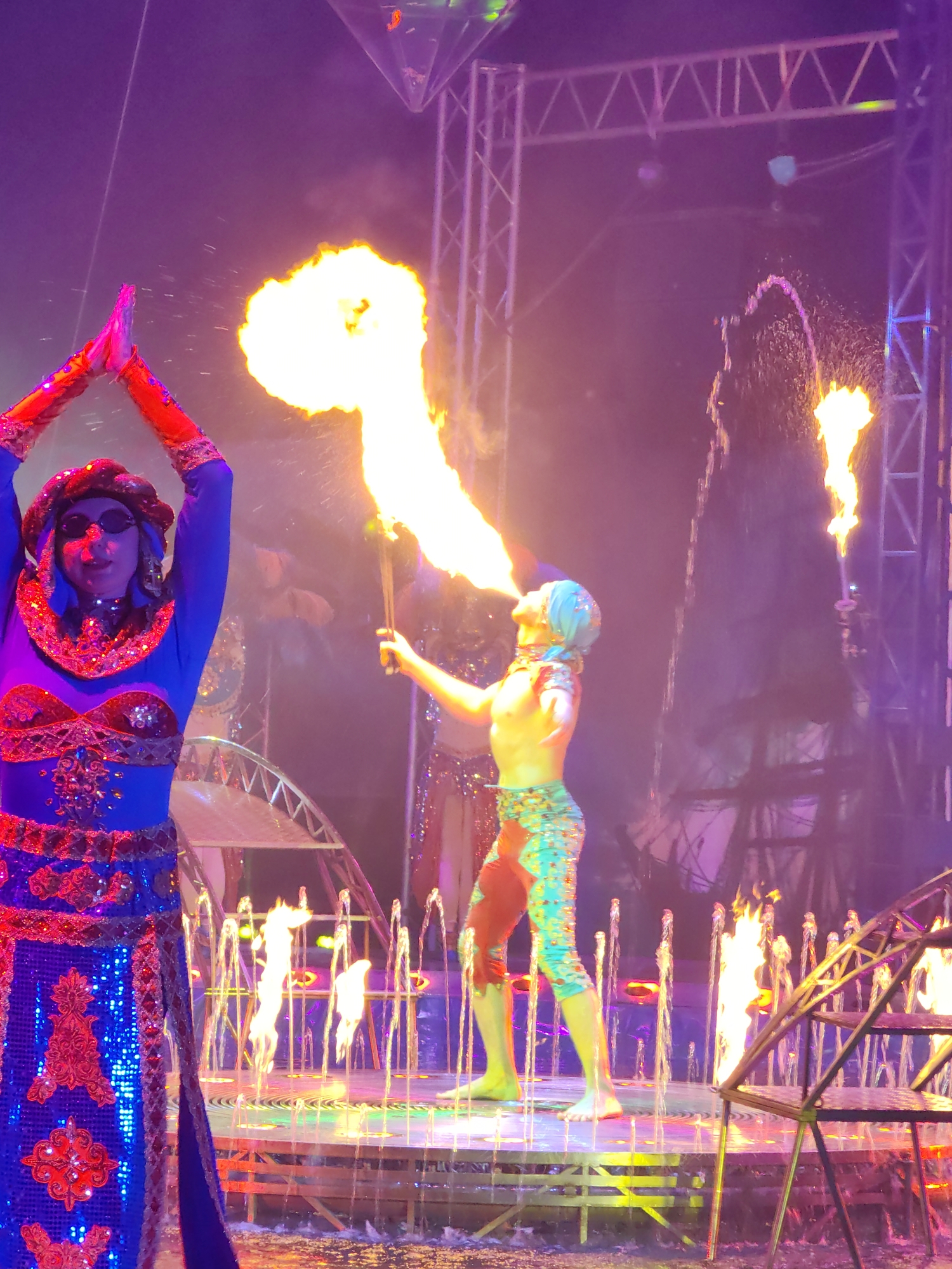 A vivid, theatrical performance is underway, likely in an entertainment venue in Tver, Russia. Two figures are prominently featured. On the left, a person is wearing a blue, sequined costume with red embellishments and sunglasses, their hands raised in a gesture of performance. To the right, another figure, shirtless and wearing patterned leggings and a head covering, is blowing fire from their mouth, holding a torch. Both are positioned on a stage platform surrounded by water fountains. Additional flames are visible in the background, contributing to the dramatic atmosphere. The scene is lit with bright, multi-colored lights, creating a visually dynamic spectacle. The background includes scaffolding and other stage elements.