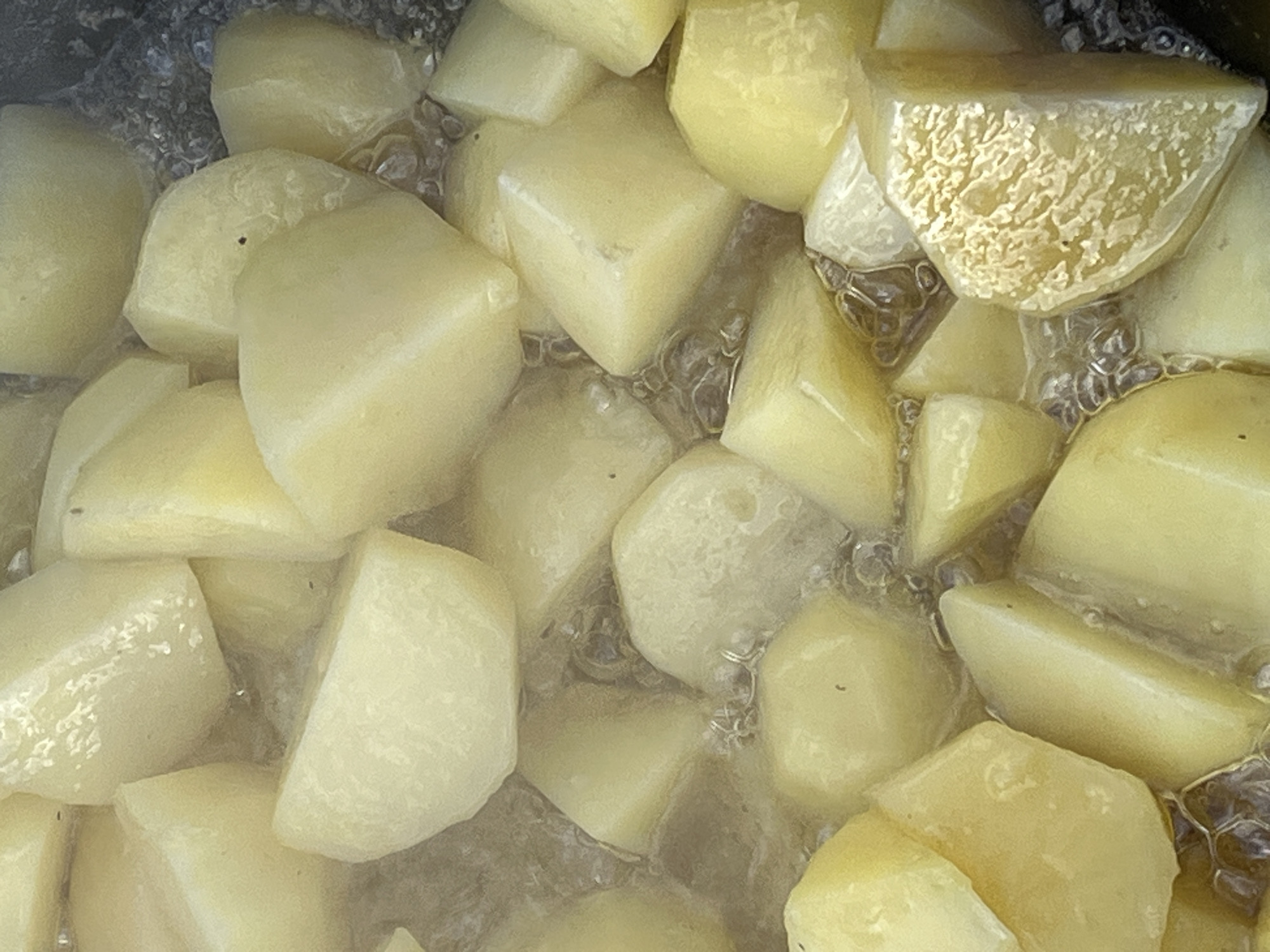 The image shows a close-up, overhead view of numerous chopped potatoes simmering in a liquid. The potatoes are light yellow and appear to be cut into irregular, bite-sized chunks. Bubbles are visible in the liquid surrounding the potatoes, indicating they are being cooked. The environment is a cooking pot, with dark, out-of-focus edges suggesting the pot itself. There are no people or specific city landmarks visible in the image. The scene is a simple, domestic cooking process.