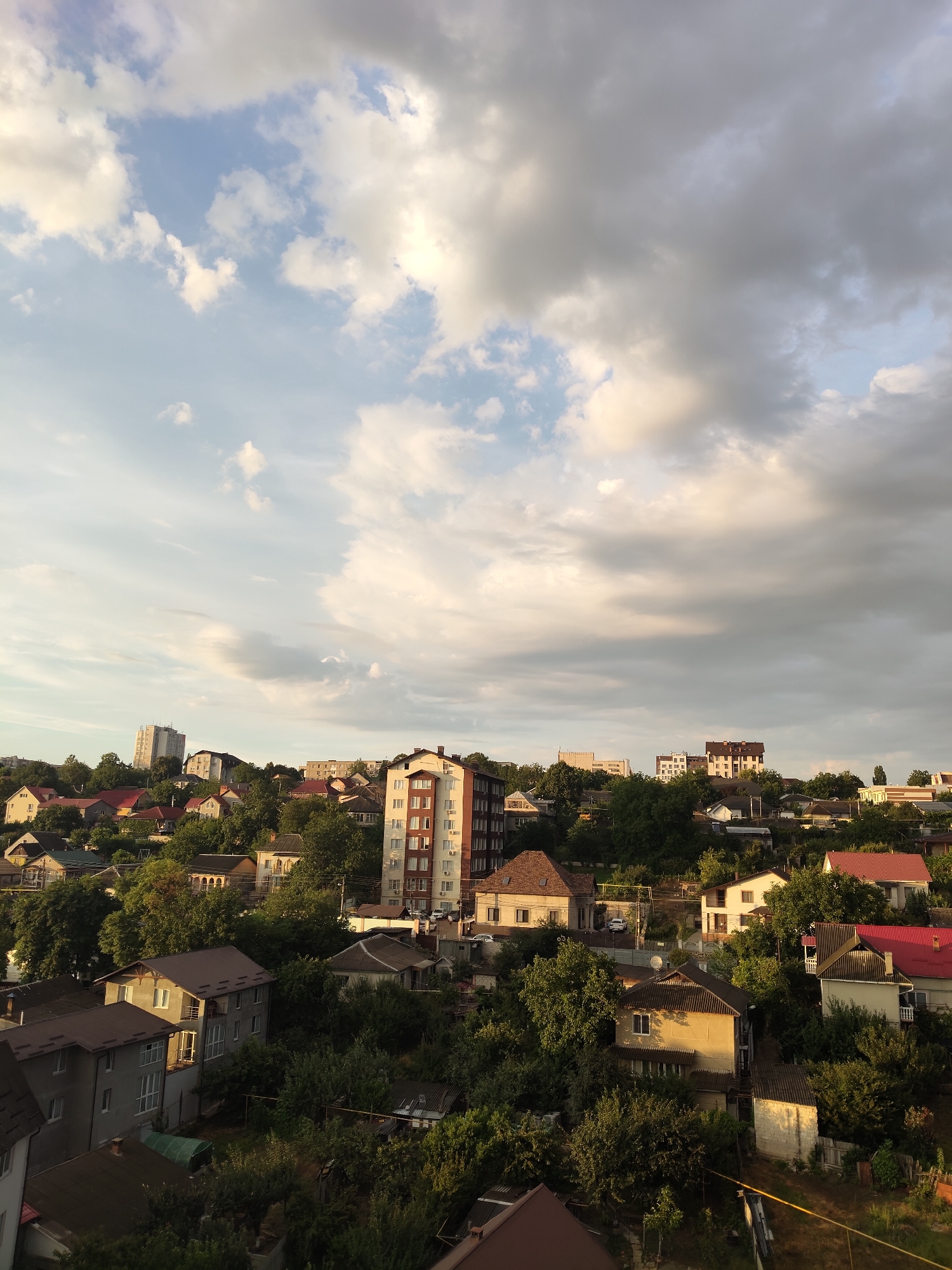 The image is a medium-angle, high-view, exterior shot of a hillside residential area in Durlești, Moldova, taken during the day. The sky is partly cloudy with a mix of white and gray clouds, some illuminated by sunlight. The foreground shows the roofs and upper floors of several houses and apartment buildings, some of which appear to be older with reddish-brown or grey roofs, while others are more modern multi-story structures. Dense green trees and foliage are interspersed throughout the residential landscape, creating a natural, somewhat overgrown feel. The architecture varies from single-family homes with yards to taller apartment buildings. The overall impression is of a quiet, established neighborhood on a slope, with the soft, diffused light suggesting it might be late afternoon or early evening. No people are visible in the frame.