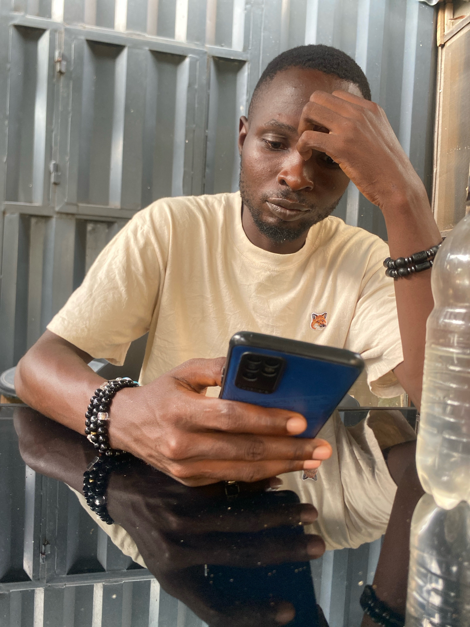 In a studio or indoor setting in Barden Yana, Nigeria, a Black male is seated at a dark, reflective table. He is wearing a light-colored t-shirt with a small fox logo on the left chest and several beaded bracelets on his left wrist. His right hand is raised to his forehead, and he is looking down at a blue smartphone he holds in his left hand. To the right of the frame, a partially visible plastic water bottle is present. The background consists of a corrugated metal wall.