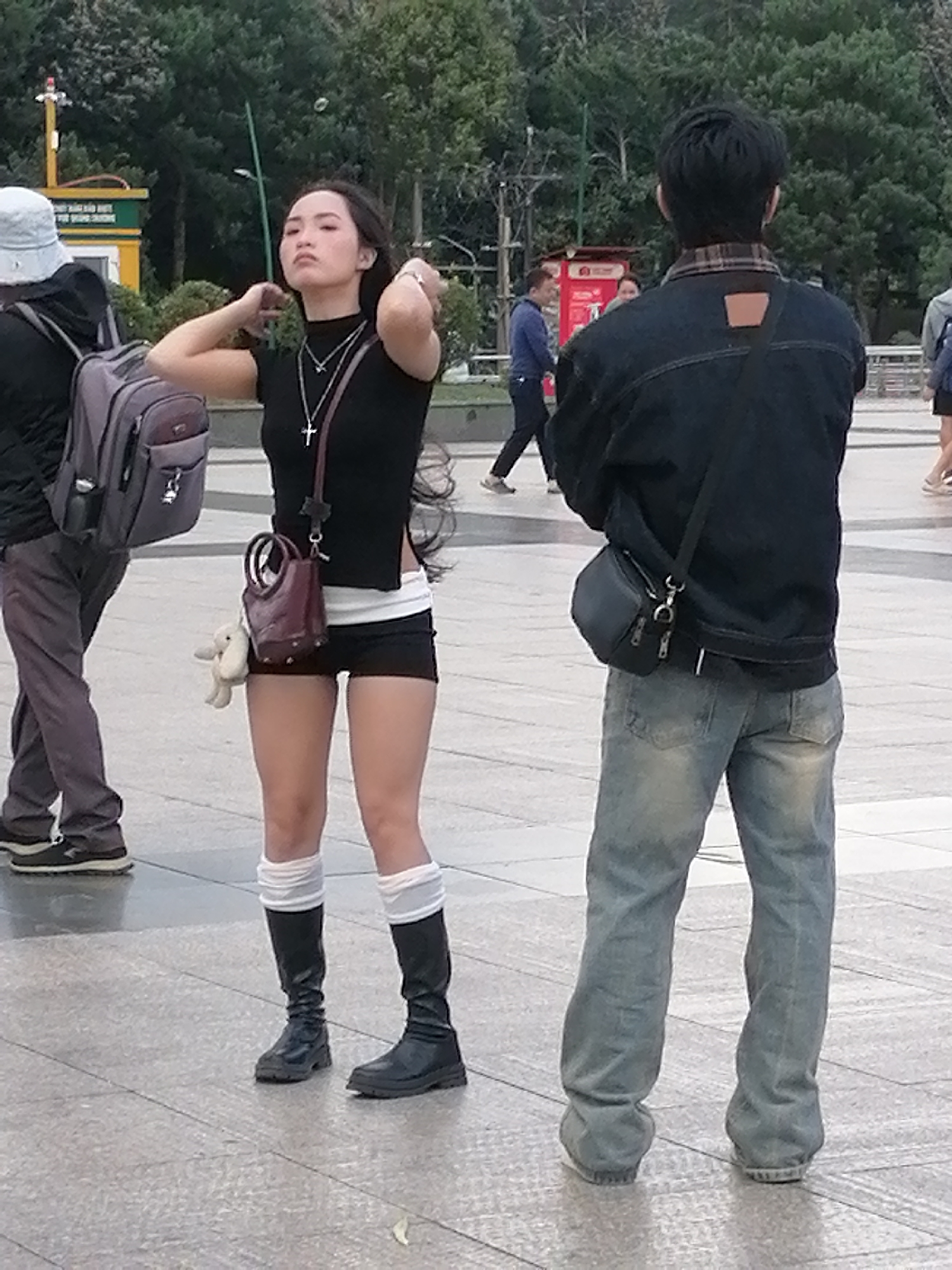 In Da Lat, Vietnam, a young woman stands facing forward, her hands touching her hair. She wears a black crop top, white briefs, black shorts, and tall black boots with white socks. A small brown purse hangs from her right shoulder. To her left, a person wearing a light-colored hat and backpack stands, partially obscured. To her right, a man in a denim jacket and light blue jeans faces away from the camera. The ground is paved with square tiles, and trees are visible in the background. The overall scene appears to be a public outdoor area.