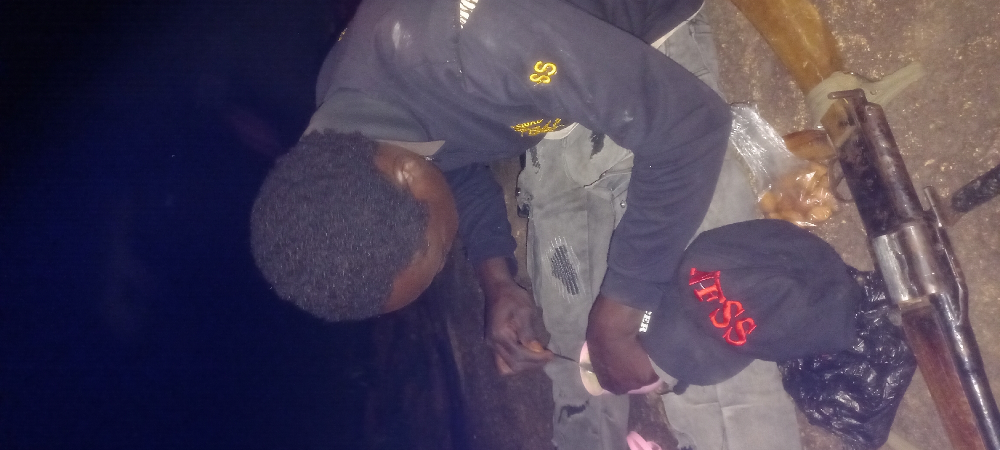 In Kwang, Nigeria, a person is captured in a dimly lit outdoor setting, possibly at night, as suggested by the dark background and artificial lighting. The individual, wearing a dark blue sweatshirt with yellow lettering on the sleeve, is seen hunched over, engaged in an activity that involves a small cup or container and a spoon-like object in their hands. The person appears to be mixing something in the container.
On the ground near the person, a black baseball cap with red embroidery of the letters "SS" is visible. Also on the ground is a black plastic bag and a portion of what appears to be a firearm, with its metallic components and wooden stock in view. The overall scene suggests a moment of preparation or engagement with unspecified substances in a somewhat clandestine environment. The ground appears to be a rough, uneven surface, possibly dirt or gravel.