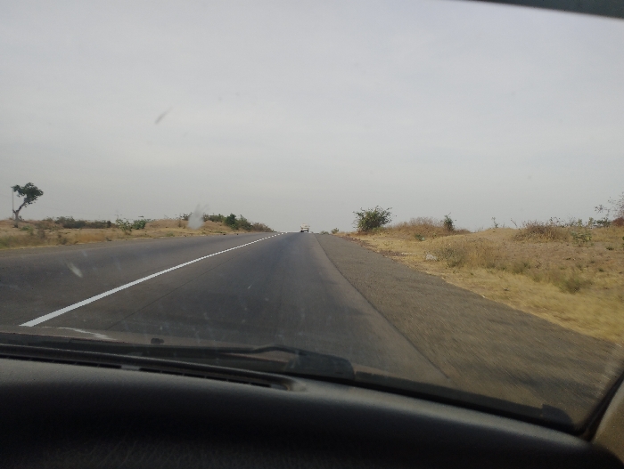 The image shows a view from the driver's seat of a vehicle traveling on a two-lane paved road. The road is straight and stretches into the distance. There is a white dividing line in the center of the road. On the right side of the road, there is a grassy shoulder with scattered shrubs and trees. There is a single white vehicle in the distance, traveling in the opposite direction. The sky is overcast and grey. The location is a road in Kumo, Nigeria at coordinates (latitude: 10.14, longitude: 11.107).