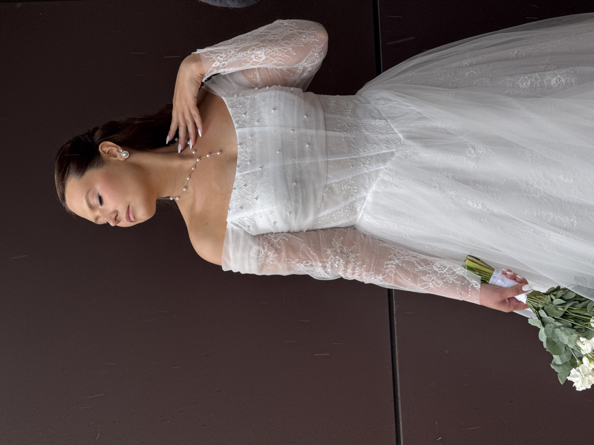 A close-up portrait shows a woman, likely a bride, positioned diagonally across the frame against a dark brown background. Her head is tilted slightly downwards to the left, and her eyes are gently closed or downcast, conveying a serene expression. She has dark hair styled in an updo and wears pearl drop earrings and a delicate pearl necklace.
The woman is adorned in a white bridal gown featuring intricate lace patterns on the sheer, long sleeves. The bodice of the dress is embellished with small, iridescent pearls and possibly sequins. Her left hand, with white-painted nails, rests gracefully near her neck and shoulder. Her right hand, mostly out of frame at the lower right, holds a bouquet consisting of white flowers, resembling carnations, and green eucalyptus foliage.
The background is uniformly dark, providing a stark contrast that highlights the white of her dress and complexion. The lighting is soft and even, emphasizing the details of her attire and jewelry. The overall mood is one of tranquility and elegance. The setting for this portrait is Chaikovsky, Russia. No discernible text is visible in the image.
