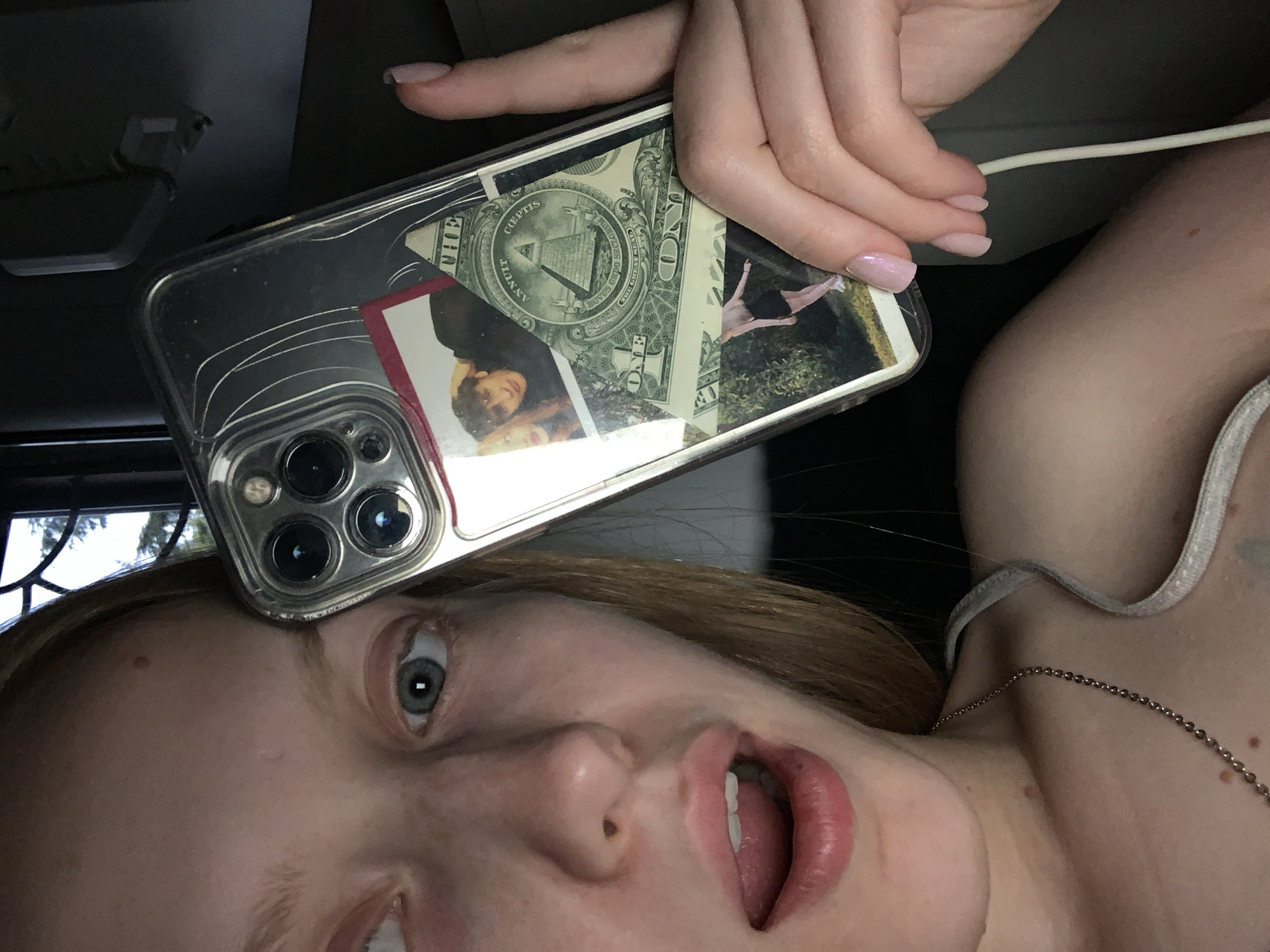 The image is a close-up selfie of a young person holding a clear phone case adorned with collaged images and a dollar bill. The person has blonde hair and blue eyes and is looking at the camera with their mouth open. The phone case features a photograph of two people, a dollar bill with the Eye of Providence, and a picture of a person in a black skirt and white shoes. The background is dark, suggesting an indoor environment, possibly a car due to the shadowy interior and a glimpse of a window with trees visible outside. The overall lighting is dim, with the flash of the camera illuminating the subject and the phone.