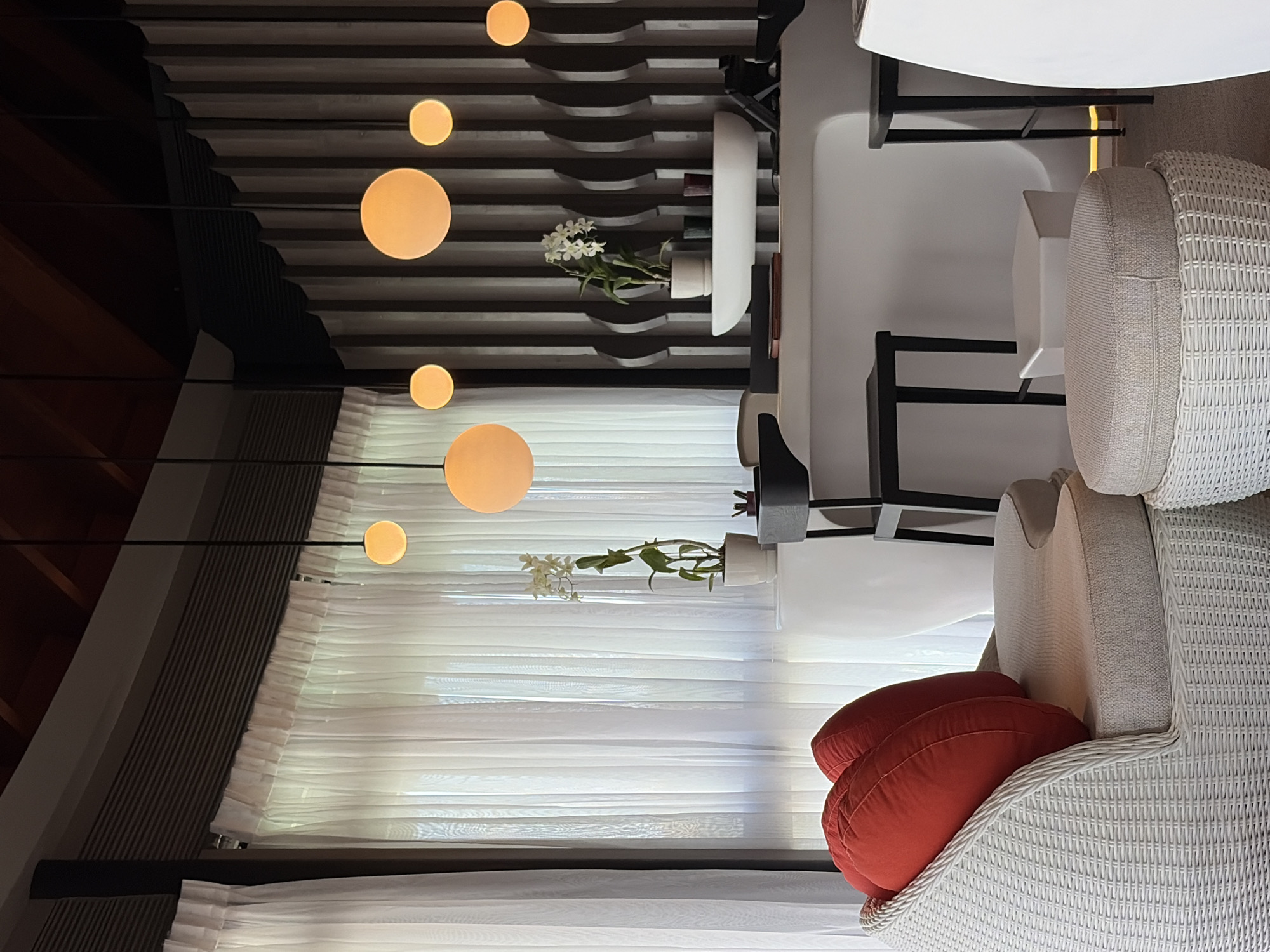 The image depicts a reception area with a white curved counter, adorned with a white bowl on which a potted white orchid sits. Behind the counter, a dark, fluted wall serves as a backdrop. Several spherical, amber-colored pendant lights hang from the ceiling, illuminating the space. In the foreground, a white wicker armchair is furnished with two large red cushions. To the right of the armchair, a round, beige ottoman rests on a white wicker base. Two black bar stools are positioned near the counter, and a white geometric ottoman is situated between them. Sheer white curtains are visible in the background, framing the view through a window. The scene is devoid of people. This image captures a serene interior setting, likely within a resort or hotel in Halaveli, Maldives.