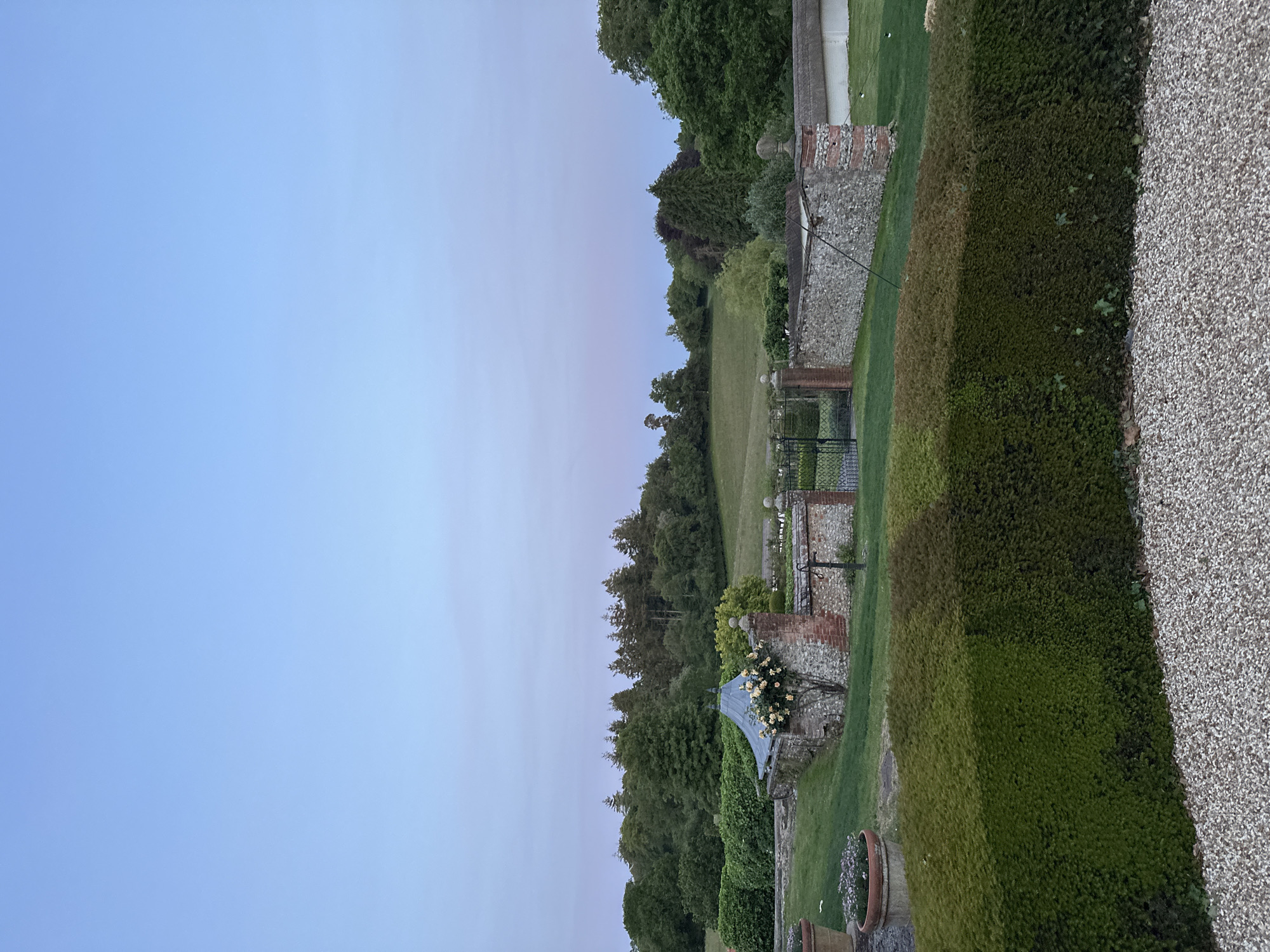 The image captures a landscape at dusk. The sky transitions from blue to a soft pink hue. Below the sky is a dense line of trees. Further down, a stone wall is partially visible, interspersed with red brick sections and green foliage. A dark metal gate is positioned within the wall. In the foreground, a tall, neatly trimmed hedge runs along the right edge. To the far right, a textured wall is visible. The scene appears to be in a garden or park setting. The location is Unknown, Unknown.