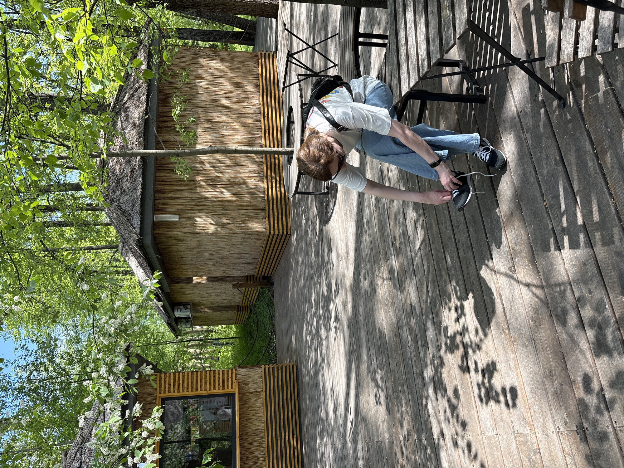 A person is sitting on a black metal chair on a wooden deck, tying their shoe. They are wearing a white t-shirt, blue jeans, and black shoes with white laces. The person is facing forward with their body bent at the waist. A black backpack is hanging on the back of the chair. The deck is next to a wooden building with a thatched roof. The area appears to be outdoors with trees and sunlight. The location is Izhevsk, Russia.