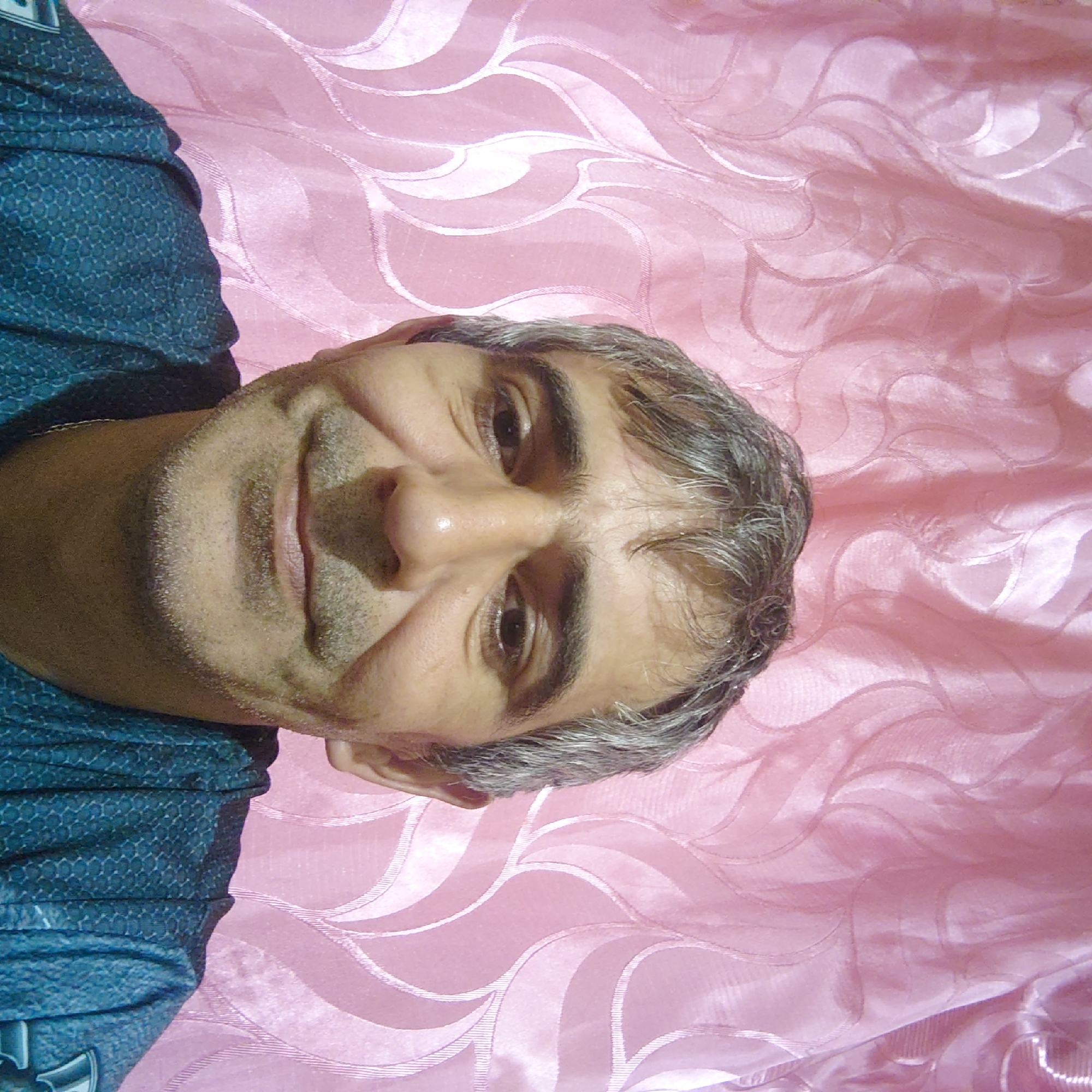 A man with graying hair and a light beard is looking into the camera. He is wearing a dark blue shirt. The background is a pink curtain with a wavy pattern. The image appears to be a selfie taken indoors. There is no indication of the specific location within Sokolohirsk, Ukraine, beyond the stated fact that it is in that city.