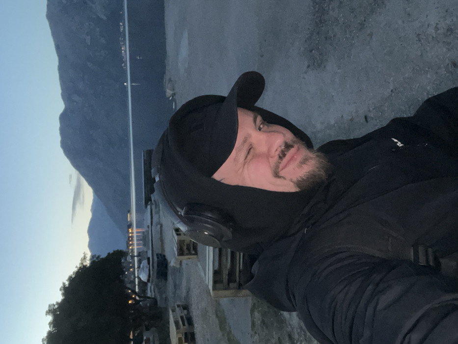 An outdoor, eye-level, slightly off-center portrait captures a male subject at dusk in Bogdašići, Montenegro. The male, positioned in the foreground, is winking with his right eye (viewer's left) and exhibiting a slight smile. He has a short beard and mustache, and a silver-colored hoop nose ring is visible on his left nostril. He is dressed in multiple layers of black clothing, including a baseball cap, a hooded sweatshirt, and a padded jacket. Black over-ear headphones are worn over the hood. A dark strap, possibly from a backpack, is visible over his left shoulder.
The background features a calm body of water, consistent with a bay or fjord, reflecting the ambient twilight sky. Distant mountains rise from the water's edge, silhouetted against the horizon. Lights from a distant settlement are visible on the far side of the water, creating reflections on its surface. The immediate shoreline consists of gravel or coarse dirt. To the left, dark trees with visible foliage are present. Several stacks of wooden pallets are discernible on the shore behind the subject, along with indistinct objects resembling small boats or covered structures near the water's edge. The overall lighting is dim, indicating late evening or early night.