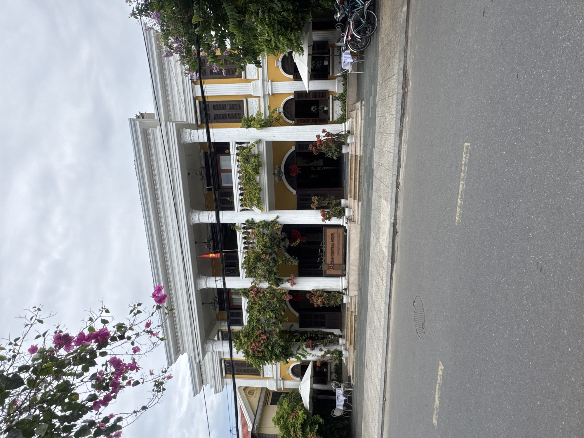 The image depicts a yellow, colonial-style building with white columns, likely a hotel or business, on a street in Hội An Ward, Vietnam. The building has two stories with dark shutters on the windows and is adorned with flowering vines. A Vietnamese flag hangs between the columns. There is a sign that reads "Cam Central Hotel." The street is paved and has a sidewalk. Bicycles are parked near the building. Part of a tree with purple flowers is in the foreground. The sky is overcast. There are chairs and a table near a white umbrella that appear to be part of an outdoor eating space.