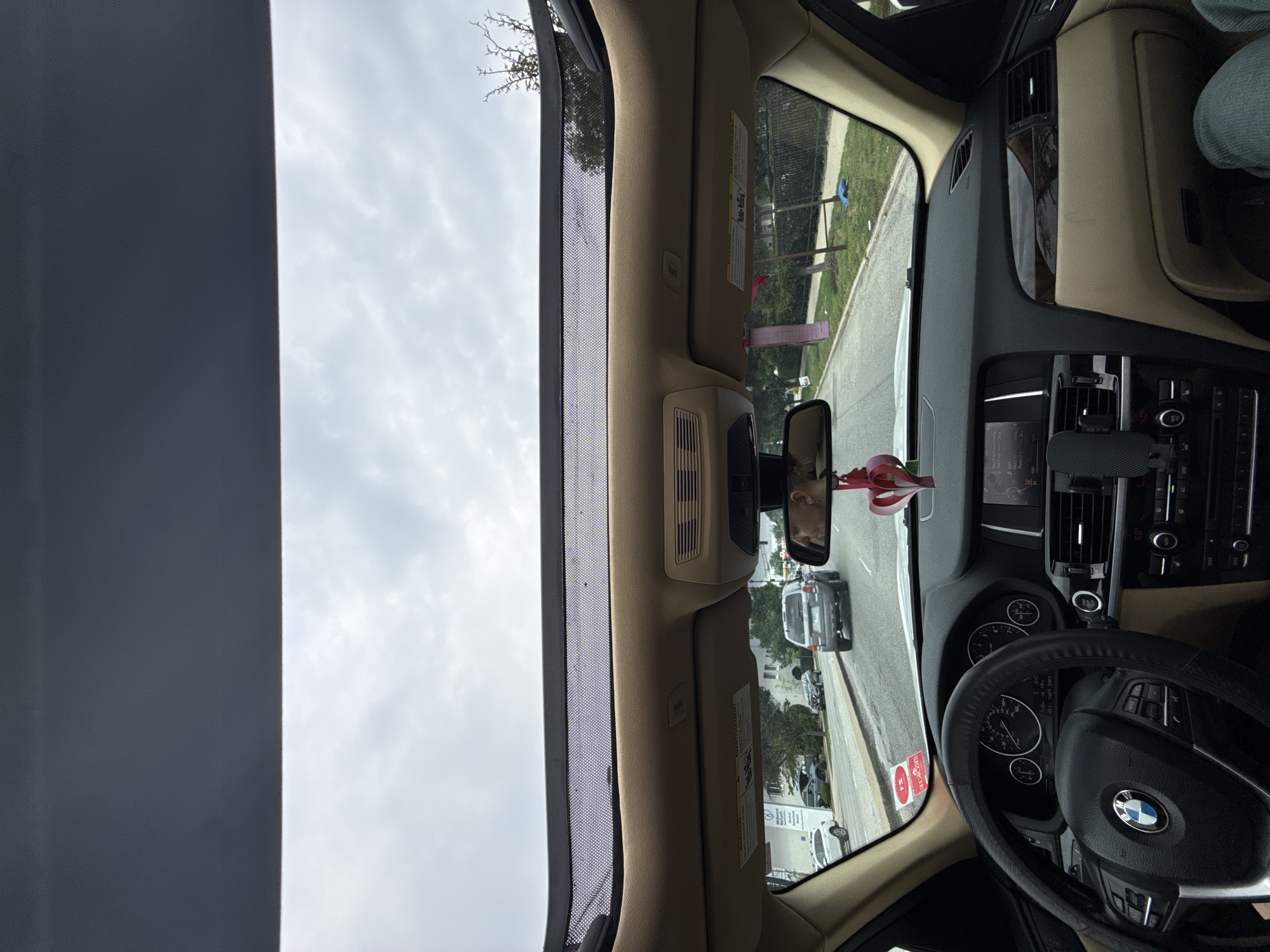 The image shows the interior of a car, looking forward through the windshield and sunroof. The car appears to be in motion, driving on a street lined with trees and buildings. A gray SUV is visible ahead in the right lane, and other cars are on the road. The dashboard of the car is visible, including the steering wheel, instrument cluster, and infotainment screen. A heart-shaped ornament is hanging from the rearview mirror. The sky is overcast. The location is Lincolnwood, Illinois, USA.