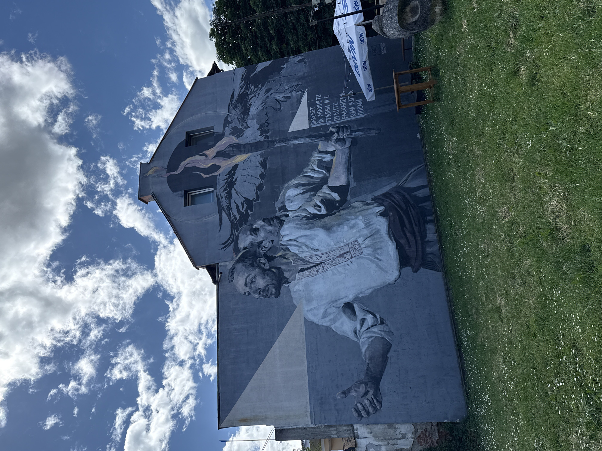 A striking mural covers the side of a building under a bright blue sky with scattered white clouds, likely during daytime. The artwork, primarily in grayscale with hints of yellow and orange for a flame, depicts a determined-looking man in traditional attire, holding a tall, flaming torch or staff aloft. Behind him, a large, powerful bird, possibly an eagle, is visible with outstretched wings, and above his shoulder, another weathered or skull-like face emerges from the shadows. The mural fills the entire side of the multi-story building, which also features two windows integrated into the design.
In the foreground to the right, a patch of vibrant green grass speckled with small white flowers stretches out. Nearby, a small wooden table and a stone object, resembling an urn or sculpture, are visible. Above, white banners with the logo "DEVIN" (a Bulgarian mineral water brand) and partially visible "ROCKILIMON" hang, suggesting a commercial or event setting. Trees are also visible in the background.
The mural includes Cyrillic text, which reads:
"ФАКЕЛЪТ НА ЗНАНИЕТО" (The Torch of Knowledge)
"ГРАБНИ И С ОРЛИТЕ БАЛКАНСКИ" (Grab and with the Balkan Eagles)
"КЪМ ВЪРХА ПОЕМИ" (Towards the Peak Set off/Undertake).
This poetic inscription, combined with the "DEVIN" branding and the provided context, places the scene in Vratsa, Bulgaria. The overall impression is one of inspiration and resilience.