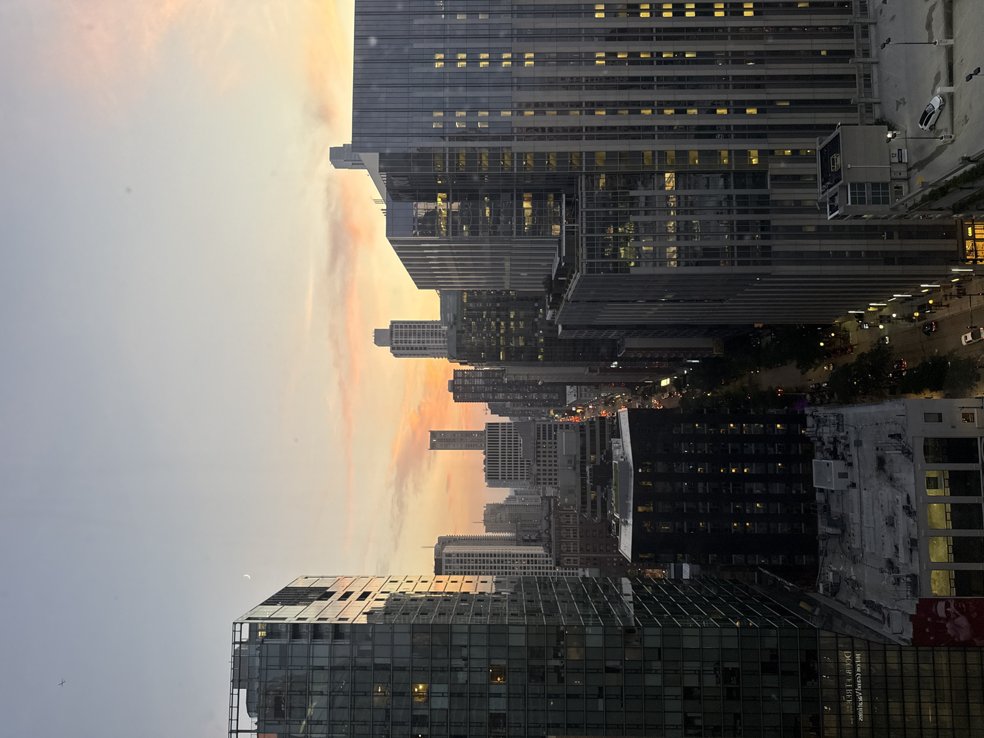 The image depicts a twilight view of a cityscape, likely Chicago, USA. The sky displays a gradient of pastel colors, transitioning from a pale blue at the top to soft pinks and oranges near the horizon, with a crescent moon visible in the upper left quadrant. Numerous skyscrapers dominate the frame, their glass facades reflecting the fading light and the illuminated windows within.
In the foreground on the left, a tall, modern building with a dark glass exterior is prominent. To its right, a street lined with trees and illuminated streetlights runs through the city. Cars are visible on the street and in what appears to be a parking structure on the right side of the image. The density of buildings and the architectural style are characteristic of a major metropolitan area. In the mid-ground and background, a complex network of buildings of varying heights and designs stretches into the distance, suggesting a bustling urban environment. The overall scene captures a moment of transition from day to night, with the city lights beginning to twinkle against the darkening sky.