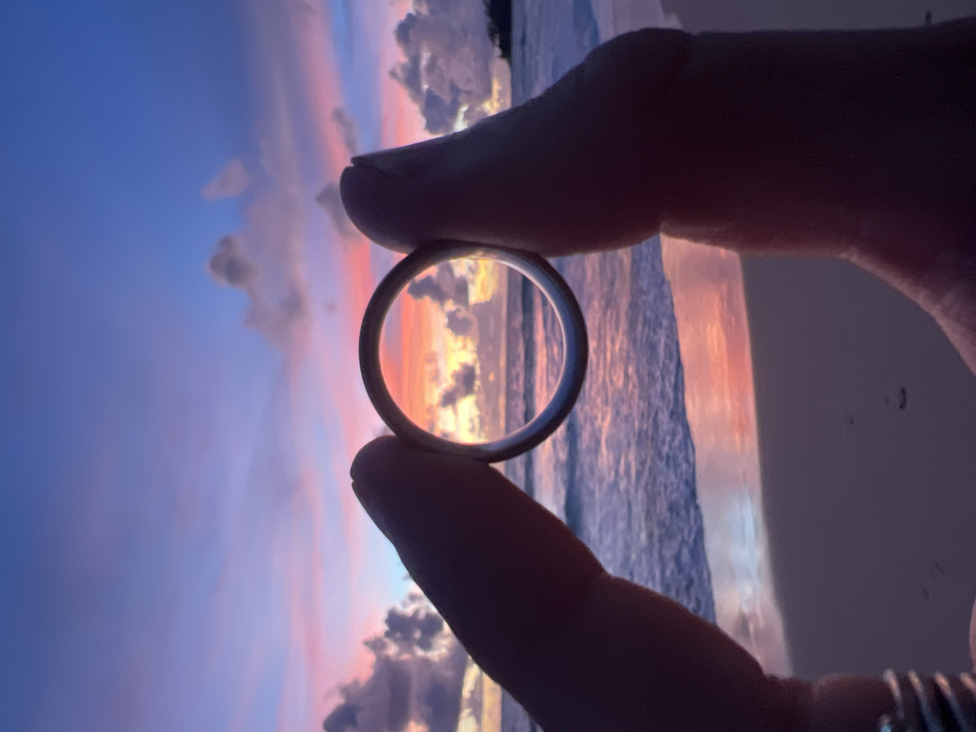 In this close-up, silhouetted shot, a person's thumb and index finger hold a ring up to frame a sunset over the ocean. The ring, a simple, metallic band, acts as a lens, focusing the viewer's attention on the vibrant sky. The sky is ablaze with the warm hues of a sunset, transitioning from a deep, clear blue at the top to soft pinks and oranges near the horizon. Wispy clouds are painted with the same warm colors, creating a dramatic and colorful backdrop.
The ocean is visible through the ring, with gentle waves lapping at the shore. The water reflects the colors of the sky, adding to the serene and picturesque scene. The foreground is dominated by the silhouetted fingers and thumb, their dark forms contrasting sharply with the bright sunset. The texture of the skin is visible, adding a human element to the otherwise natural landscape. On the index finger, several stacked rings are also visible, hinting at a personal connection to jewelry.
The time of day is clearly sunset, indicated by the intense colors in the sky and the soft light. The location is implied to be a coastal area, likely a beach, given the presence of the ocean and the sandy texture of the shoreline. The overall mood is one of tranquility and beauty, with the framing of the ring suggesting a moment of reflection or appreciation for the natural world. There is no visible text in the image. The scene evokes a sense of peace and wonder, capturing a fleeting moment of natural beauty through a simple, everyday object.