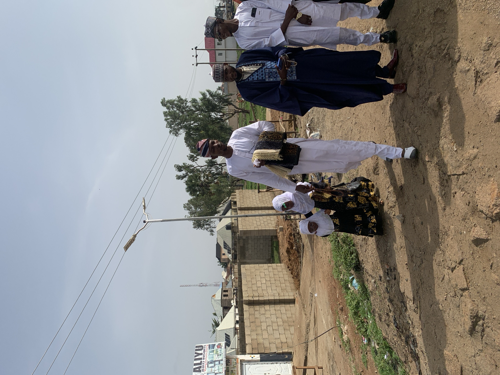The image depicts several individuals in the city of Naraguta Hausawa, Nigeria. Three men are standing on a dirt road; one in a white robe holding what appears to be folded textiles or garments, another in a dark blue robe, and a third in a white robe. To the side of the road, two women wearing white head coverings stand near a dark fabric with gold patterns, possibly clothing or a covering. A row of cinder block buildings are visible behind the group. A sign, with the name "ALIYU MULTI BUSINESS", is visible in the foreground along with a streetlight. Power lines run above the scene. The environment is dusty, and some trash and vegetation are present along the roadside.
