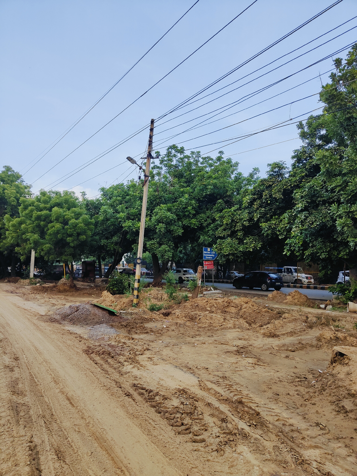 The image depicts a dirt road under construction in Gurgaon, India. The road is muddy with visible tire tracks, indicating recent vehicle activity. Piles of dirt and gravel are scattered along the roadside, suggesting ongoing landscaping or road work. Several trees line the road, providing shade. In the background, a black car is parked on the side of a paved road, which is separated from the construction area by a low barrier. A blue sign indicating "Sector 23A Community Centre" is visible on the paved road. Overhead, power lines crisscross the sky, supported by wooden poles, one of which has a streetlamp attached. The overall scene is one of urban development and infrastructure improvement.