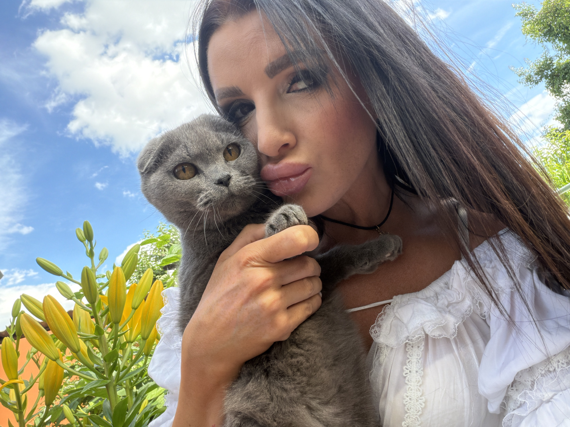 An adult female with long dark brown hair and makeup is posing for a selfie while holding a grey Scottish Fold cat. The cat has large amber eyes and is nestled in her arms. They are outdoors, with a bright blue sky and white clouds visible in the background. To the left of the frame, a cluster of yellow lily buds is in bloom. The woman is wearing a white top with ruffled lace details. The location is identified as Novaya Usman', Russia.