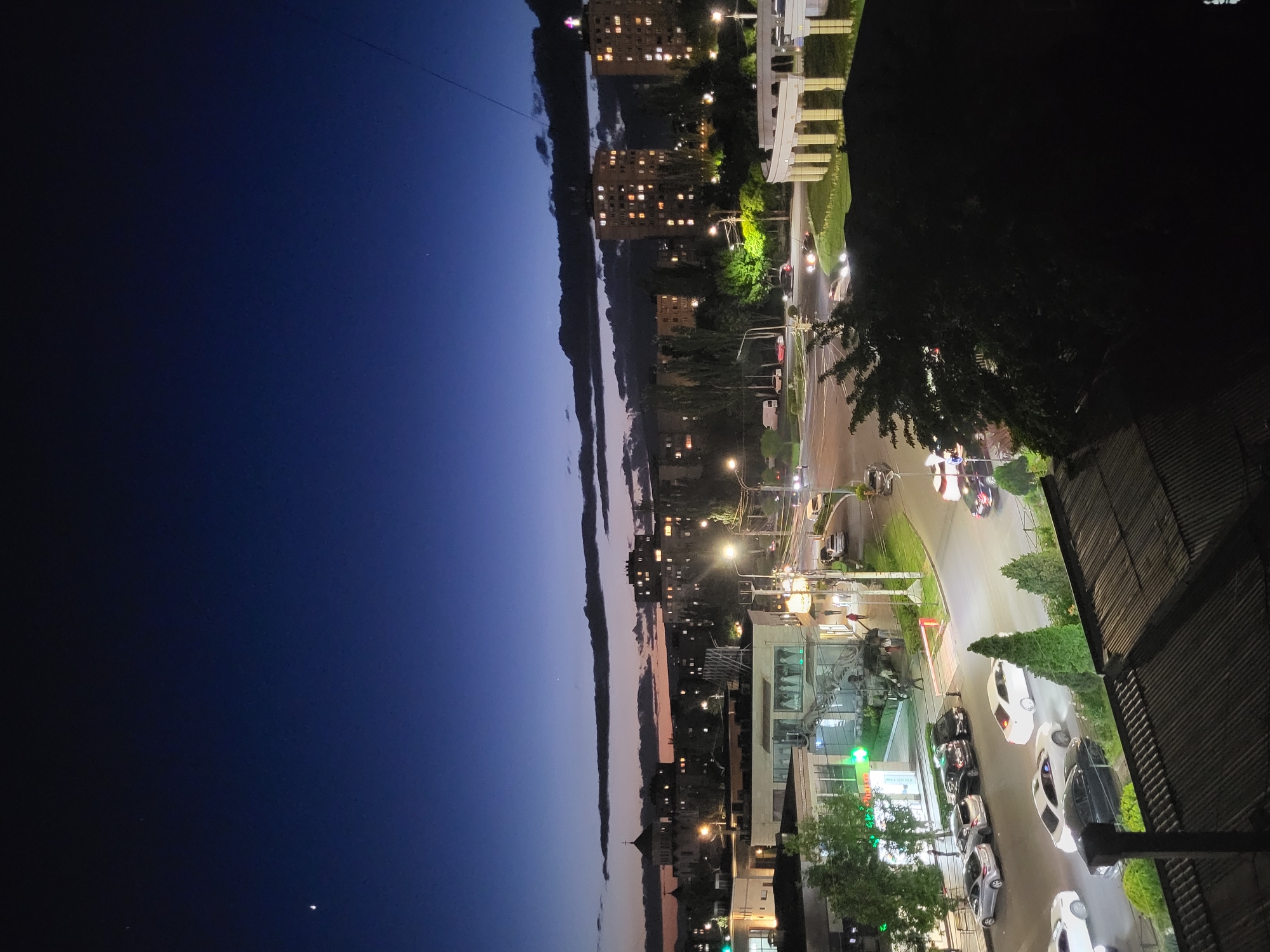 The image displays an urban cityscape at dusk or early night, captured from an elevated perspective overlooking a city street in Yerevan, Armenia. The sky dominates the upper portion of the frame, transitioning from a deep indigo near the horizon to a darker navy overhead, with a few faint stars visible. Long, narrow bands of clouds stretch across the horizon.
Below the sky, the city comes alive with artificial light. Numerous multi-story buildings, both residential and commercial, line the visible landscape, their windows glowing with interior lights. A wide street curves through the scene, bustling with traffic. Several cars are visible, some driving with their headlights and taillights creating streaks of light, while others are parked along the side. Streetlights illuminate the road and sidewalks, casting bright pools of light. Traffic lights are also visible, one clearly showing green.
Trees and other foliage are interspersed throughout the urban environment, particularly along the street and in the foreground on the right. In the lower right corner, parts of a building's roof and exterior are visible, reinforcing the high vantage point. At street level, an illuminated green sign with Armenian text is visible on a building, likely indicating a shop or a pharmacy. The overall scene conveys a calm and serene evening atmosphere in an active city.