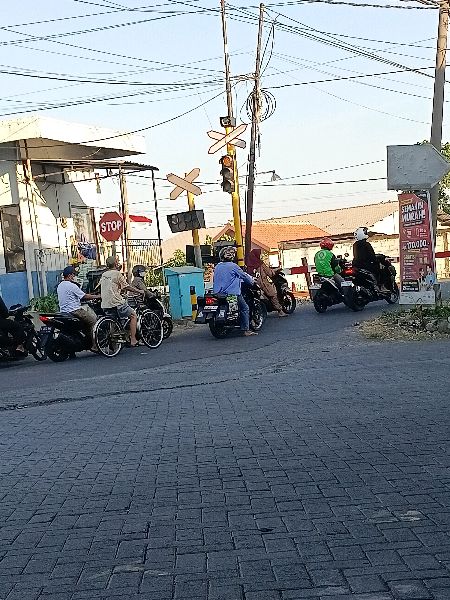 The image shows a scene at a railroad crossing in Kletek, Indonesia, with several motorcycles and their riders waiting. In the foreground, a grey paved surface with interlocking bricks is visible. Several motorcycles are lined up, some with passengers. One motorcycle has a person wearing a blue shirt and a blue cap, another has a person in a light brown shirt and a cap, and a third has a person in a dark blue shirt. Behind them, another motorcycle is visible with a rider wearing a green jacket and a red helmet, and a passenger in a dark outfit with a white helmet. Further to the right, a large billboard with Indonesian text that reads "SEMAKIN MURAH! GRATIS!" and prices is standing. A red and white striped barrier for the railroad crossing is down. A traffic signal with red and amber lights is visible, along with railroad crossing signs. A building with a white facade and a blue section is on the left. Overhead, a complex network of power lines stretches across the frame. The sky is clear and blue.