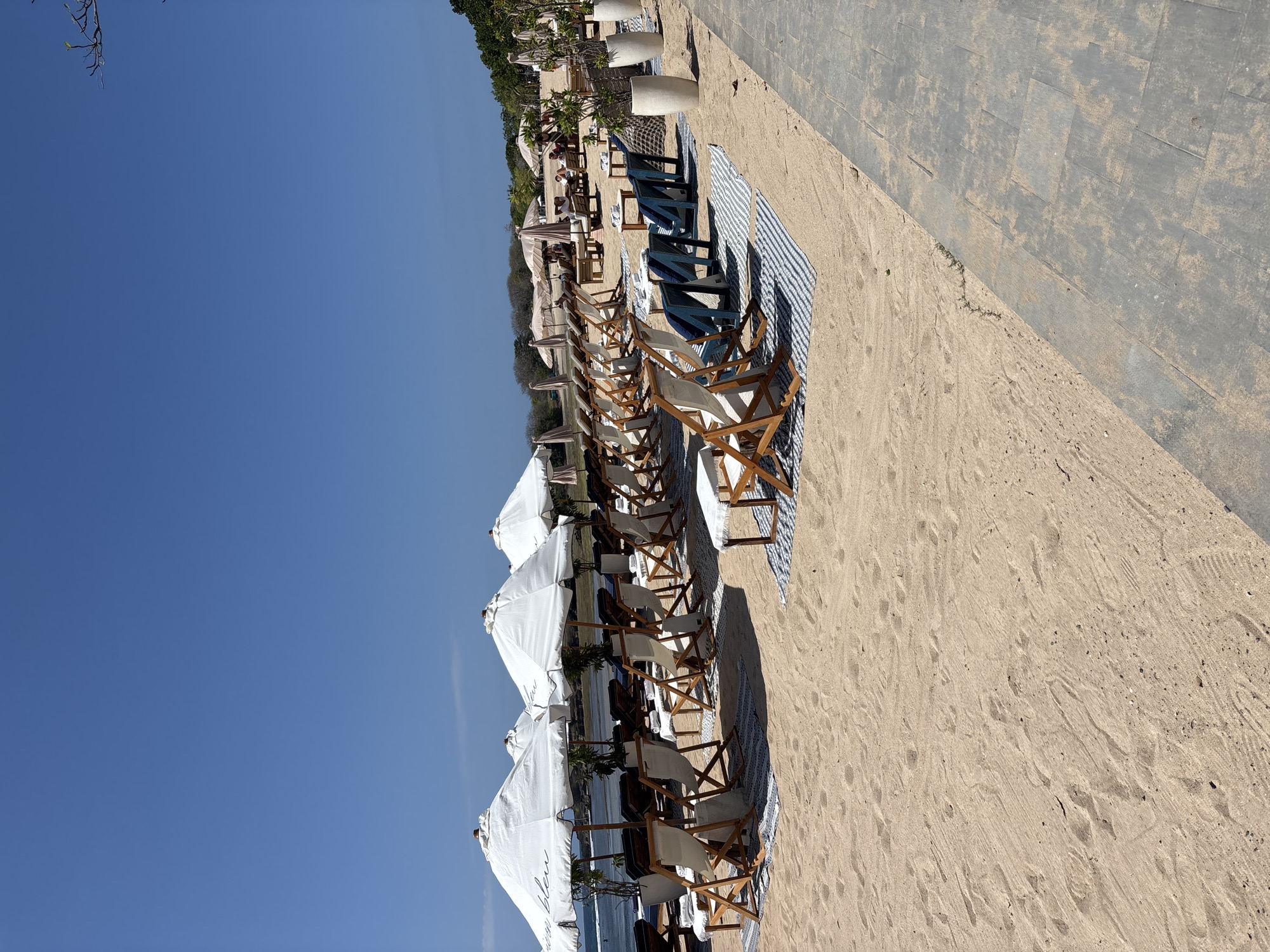 This image depicts a serene beach scene in Kuta Selatan, Indonesia, under a clear blue sky. The foreground features pristine sand with footprints, leading to a neatly arranged collection of white canvas deck chairs and tables, each placed on striped blue and white mats. Behind these, several large white umbrellas provide shade, with one displaying the text "Ocean Bleu." Further back, more deck chairs and tables are visible, some occupied by individuals. In the distance, a line of thatched-roof structures and lush greenery can be seen. The scene suggests a well-maintained beach resort or cafe offering a relaxing ambiance for its patrons.