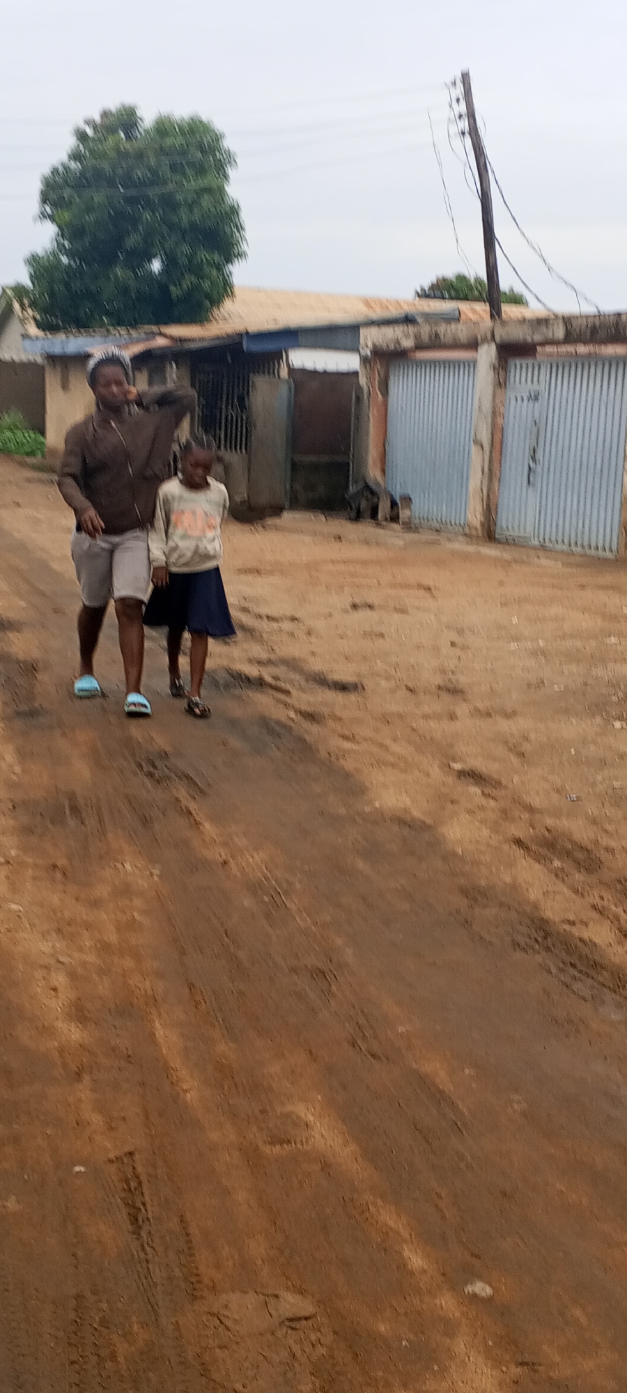 Two individuals, possibly a mother and child, walk along a dirt road in Sabon Tasha, Nigeria. The older individual, on the left, wears a brown jacket, light-colored shorts, and light-blue sandals. She is gesturing towards her head. The child, on the right, wears a light-colored sweater and a blue skirt with sandals. In the background are structures including a building with two blue corrugated steel doors and a tree. An electrical pole is on the right. The road is dirt and has tire tracks. The sky is overcast.
