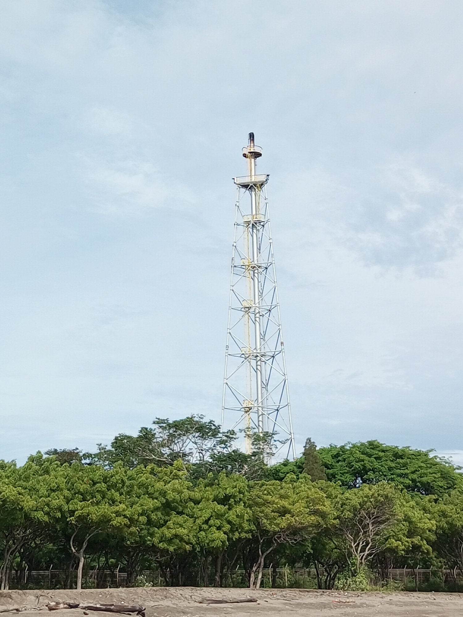 An outdoor, wide-angle, eye-level shot captures a tall, white, lattice tower with a dark, smoking pipe at its peak, set against a cloudy, pale blue sky. The tower is the dominant feature, situated slightly right of center. In the foreground, a dense line of lush green trees with visible trunks and branches stretches across the lower half of the image, creating a natural barrier. Behind the trees, a faint hint of fencing is visible, suggesting a boundary. The ground in the immediate foreground appears to be dirt or sand, with a few scattered pieces of driftwood. The overall scene is industrial yet natural, with the starkness of the tower contrasting with the vibrant greenery.