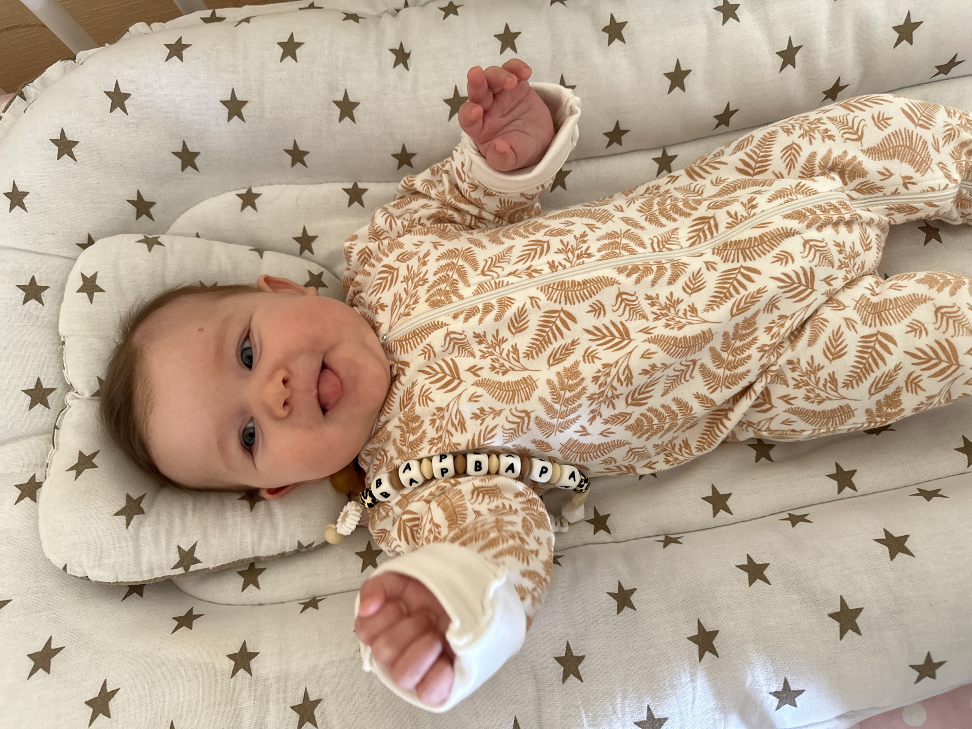 A baby wearing a printed jumpsuit lies on a star-patterned baby lounger. The baby is light-skinned with blue eyes and is sticking out their tongue. The baby is wearing a pacifier clip with alphabet beads. The lounger is placed inside a crib with a wooden rail visible in the top left. The background is a white fabric with brown stars. There is no indication of the location being Chaikovsky, Russia in the image.