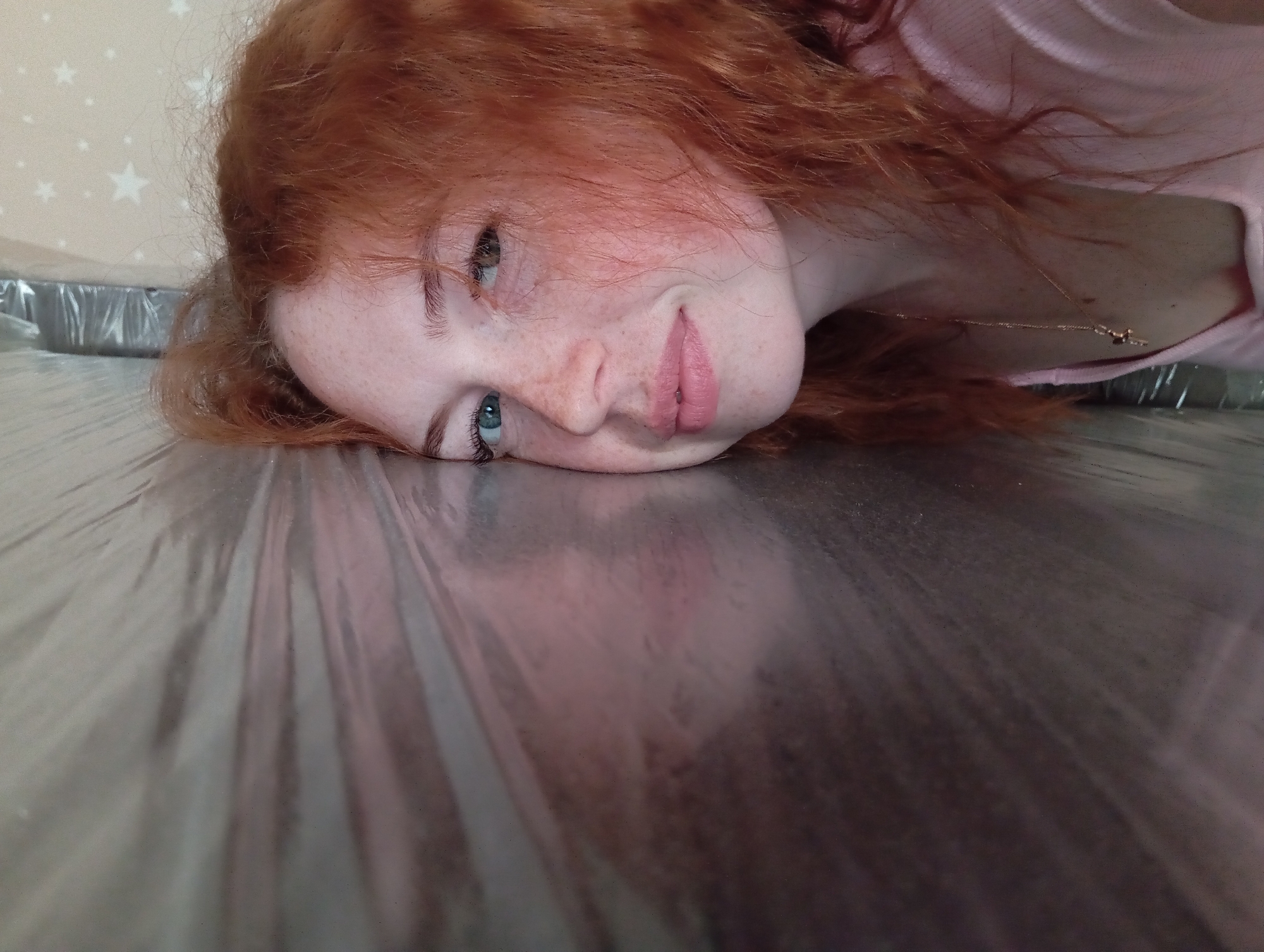 A fair-skinned young woman with freckles and reddish-orange curly hair is lying down with her head on a reflective, possibly metallic, surface. Her head is tilted towards the camera, showing her face, including her blue eyes, nose, and pink lips. She is wearing a light pink top. A delicate gold necklace is visible on her neck. The background behind her head features a light-colored wall with small white star patterns. A metallic-looking container or tray is partially visible in the top left corner behind her head. The location, Yurgamysh, Russia, is unrelated to the image content.