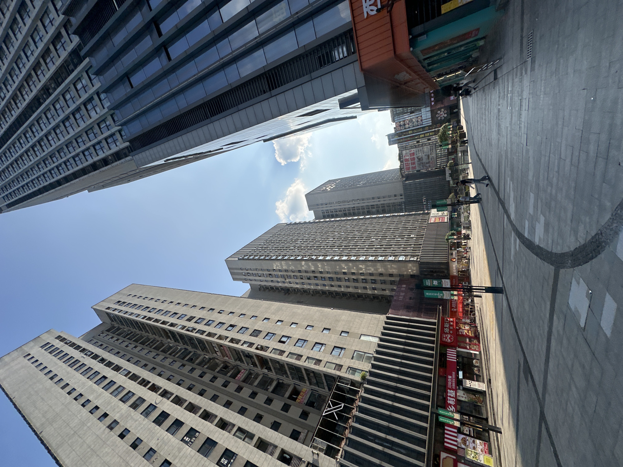 This is an outdoor, low-angle, wide shot of a street in the Chongqing High-tech Zone, China. The scene is dominated by tall, modern buildings on either side of a paved walkway. On the left, a light-colored, multi-story residential building with a grid of windows is visible. To its right, a dark, reflective glass skyscraper towers, its facade broken by horizontal bands. The right side of the street features a lower, orange-roofed building with storefronts and another tall, glass-fronted skyscraper behind it. The walkway is paved with light-colored tiles and features dark lines, with a curved section on the right. Several people are walking on the walkway, appearing as small figures in the distance. The sky above is clear blue with a few white clouds, and the sun casts shadows across the scene, indicating it is daytime. The overall impression is one of a busy, modern urban environment.