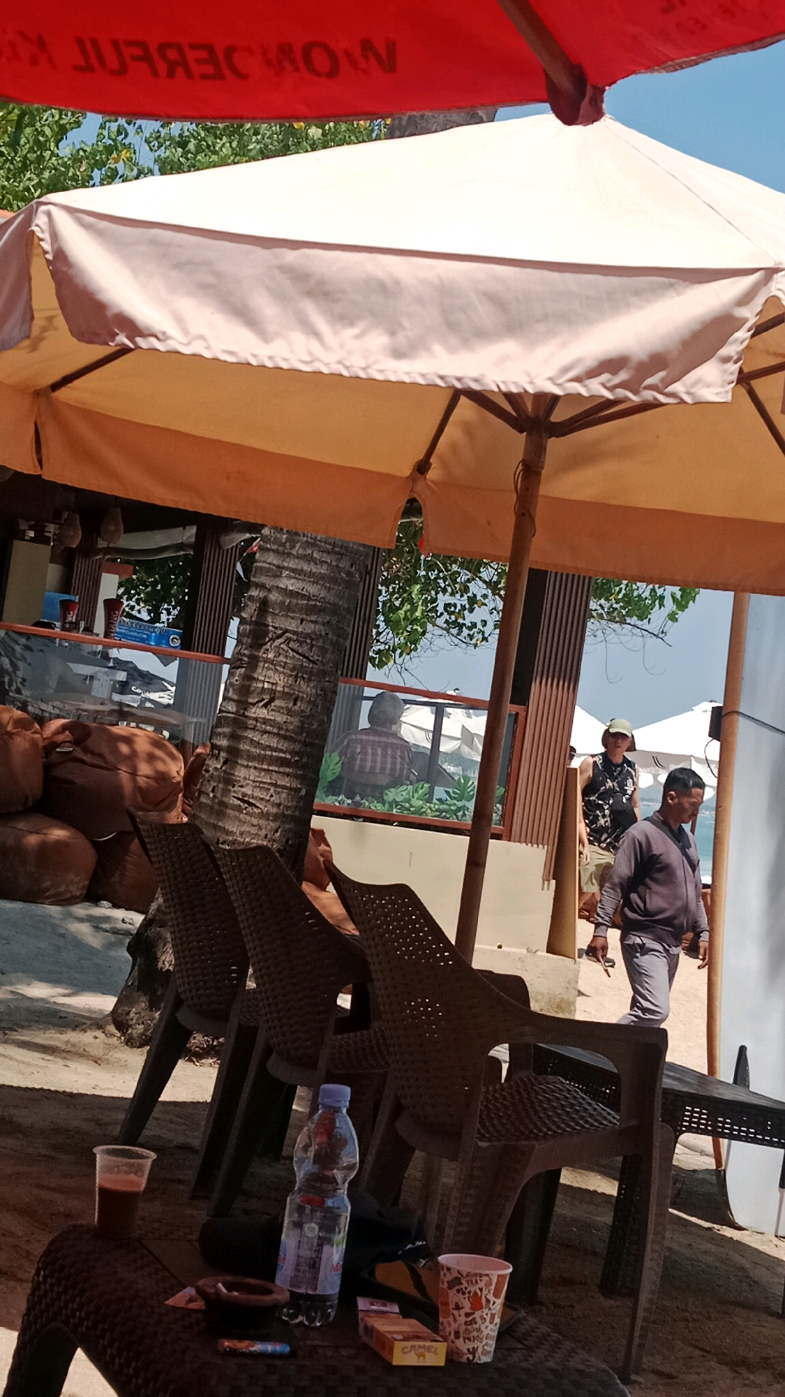 In Kuta, Indonesia, an outdoor seating area is set up on a sandy beach, shaded by a large beige umbrella. Two men are visible in the background. One, wearing a black and white sleeveless shirt and a green hat, walks towards the right. Another man in a grey jacket and pants walks to the right, following behind the first. In the foreground, a brown plastic table holds a clear plastic cup with a dark liquid, a bottle of water, an ashtray with a cigarette, a pack of Camel cigarettes, and a disposable cup with a patterned design. Behind the table, three brown plastic chairs are arranged. A portion of a palm tree trunk is visible on the left side of the image. The scene appears to be at a beachside establishment.