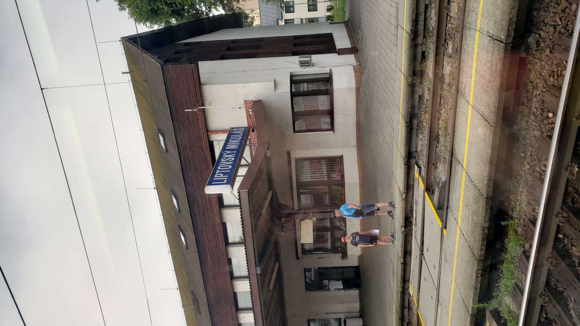 The image shows a train station platform in Liptovský Mikuláš, Slovakia. A sign on the building reads "LIPTOVSKÝ MIKULÁŠ". Two people, an adult male and a young boy, are standing on the platform facing the camera. The adult is wearing a blue t-shirt, denim shorts, and a black crossbody bag. The boy is wearing a dark t-shirt with an "Emirates" logo, dark shorts, and a red baseball cap. They are standing near the entrance of the station building, which has a covered walkway. Train tracks are visible in the foreground, separated from the platform by a concrete barrier with yellow lines. The sky is overcast.