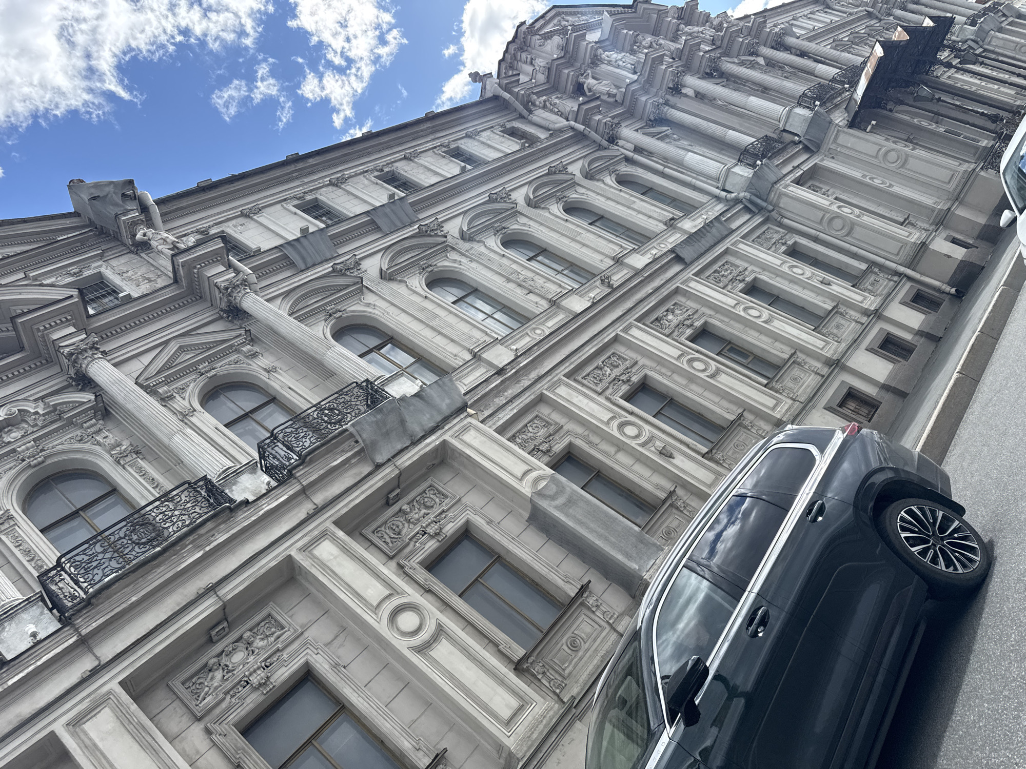 The image shows a corner of a gray, ornately decorated building in Saint Petersburg, Russia, seen from a low angle. The architectural style includes columns, arched windows, balconies with decorative wrought iron, and elaborate facade details. A dark-colored vehicle is parked on the right side of the frame, partially obscuring the lower section of the building. The sky above is blue with scattered white clouds.