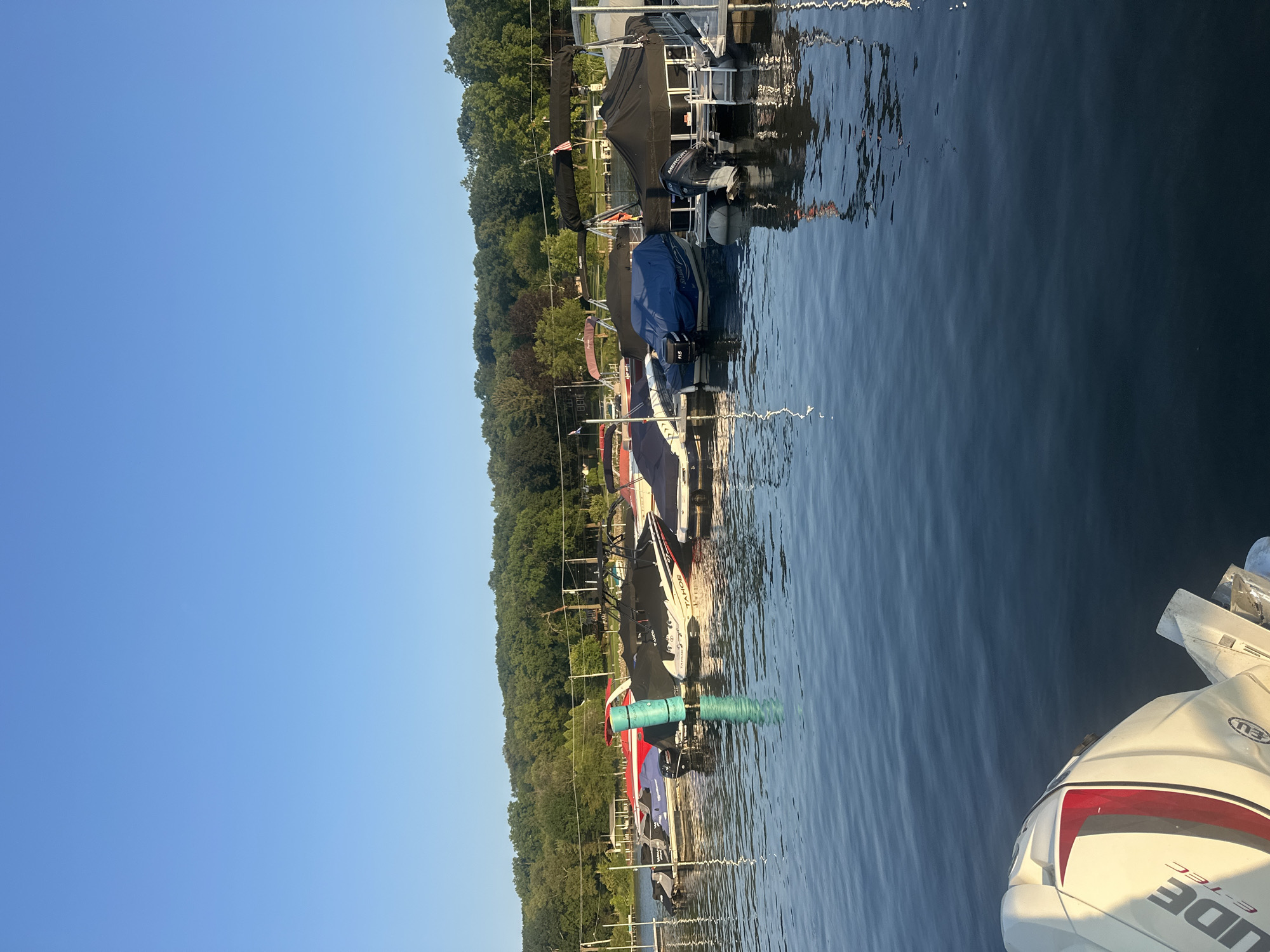 An aerial view depicts a marina on a clear day with a serene lake. The lake is populated with various boats moored along a dock, most of which are covered with black tarps. The boats range in size and style, including several pontoon boats and what appear to be speedboats. The water's surface reflects the blue sky and the boats. A lush, green forest line borders the far side of the lake, with a clear blue sky overhead. The foreground features the back of a white outboard motor, suggesting the perspective is from another boat. The location is identified as Algonquin, United States of America.