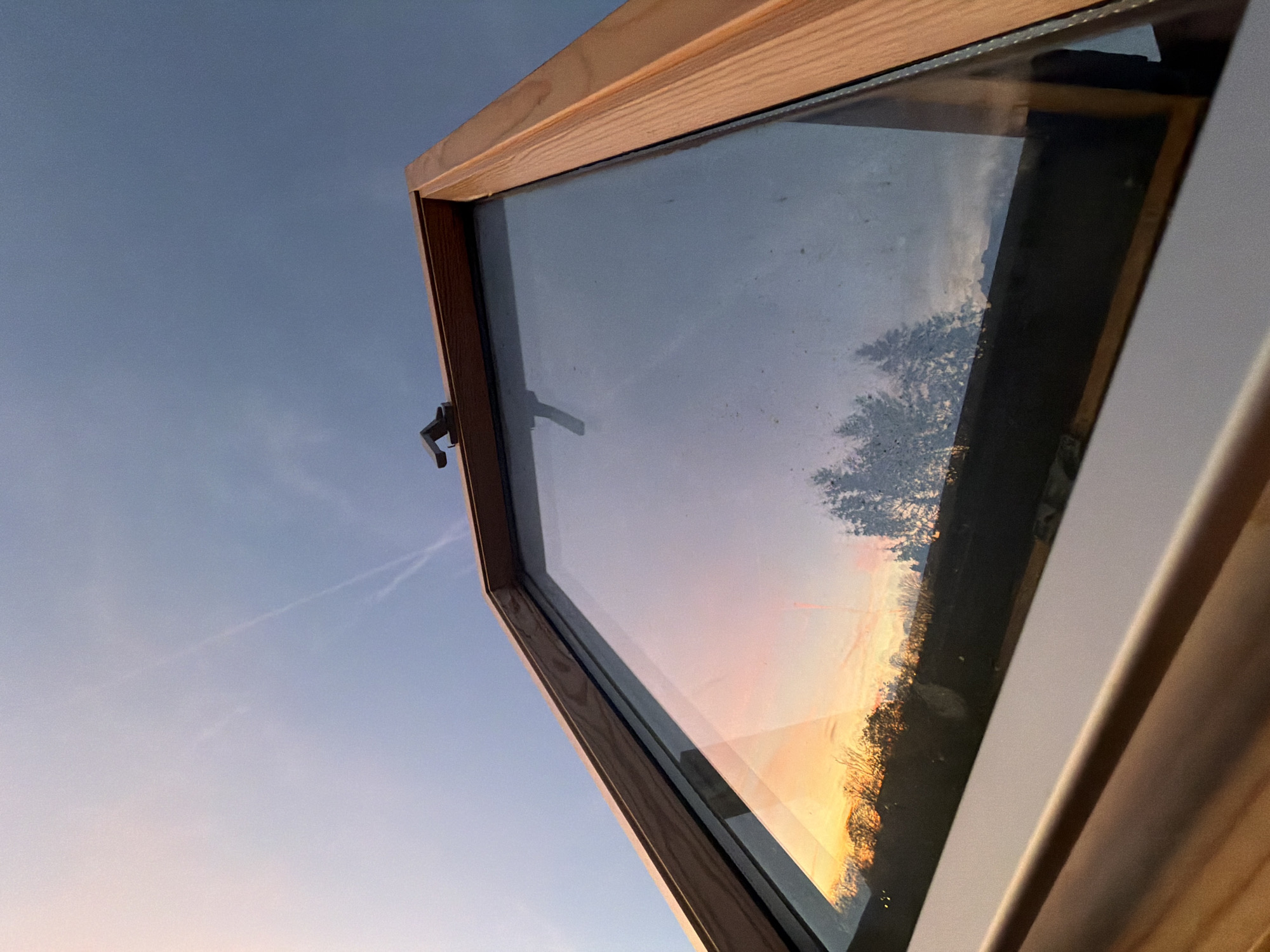 An upward-angled shot displays an open window in the foreground, reflecting a vibrant sunset and tree silhouettes in its glass. The window frame is made of light-colored wood and white trim. In the background, the sky transitions from a pale blue to a hint of pink at the horizon. A contrail bisects the upper left corner of the frame. The photo does not contain any people, actions or interactions. The location is not discernible from the visual elements presented.