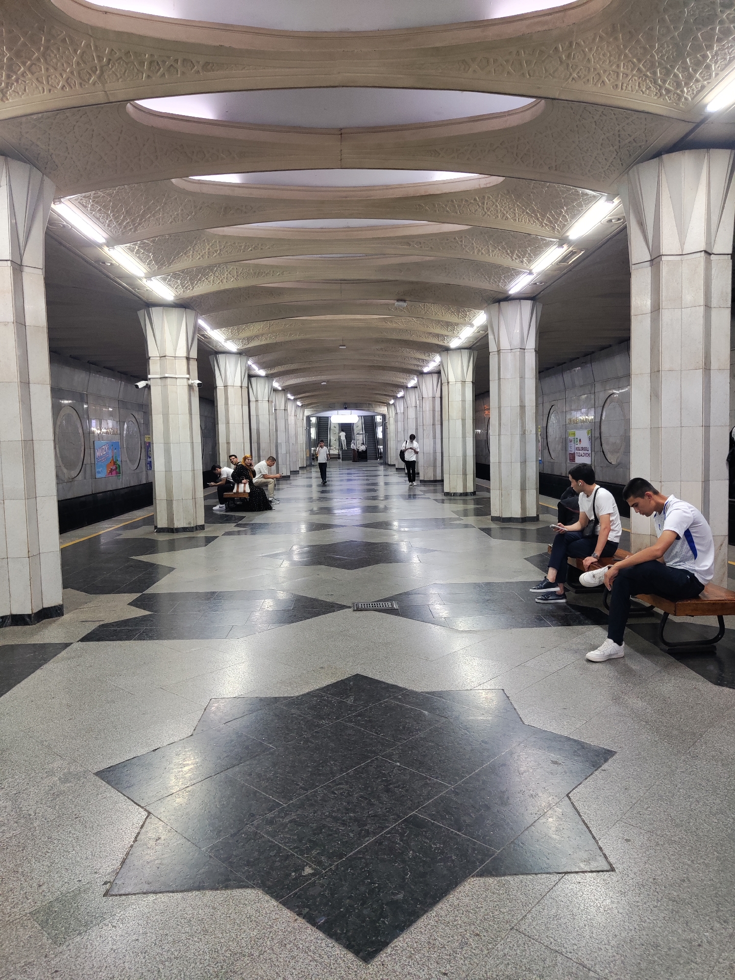 This is a photograph of a subway station in Tashkent, Uzbekistan. The station has a high, arched ceiling with decorative geometric patterns and integrated lighting. The walls are clad in marble-like tiles, and large, fluted columns support the ceiling. The floor is paved with a patterned mosaic of light and dark grey granite tiles, featuring large starburst shapes.
In the foreground, two young men are sitting on a wooden bench on the right side of the platform. The man on the left is wearing a white t-shirt and dark pants, holding a mobile phone and wearing earbuds. The man on the right is also wearing a white t-shirt and dark pants, with one leg crossed over the other. In the middle ground, a group of people are sitting on a bench on the left side of the platform. Further down the platform, a few individuals are standing and walking. In the background, escalators are visible, leading upwards. Advertising posters are displayed on the walls along the platform. The overall impression is of a clean, well-maintained, and architecturally distinctive public space.
