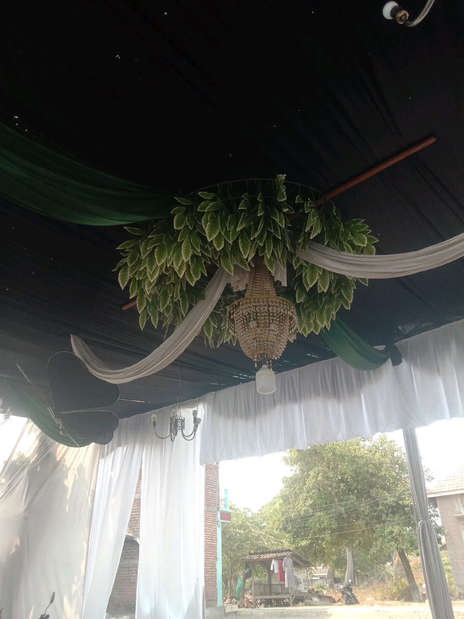 The image captures an outdoor event setup, likely for a wedding or celebration, with a dark fabric ceiling adorned with artificial greenery and a crystal chandelier. White and green draped fabrics are also incorporated into the ceiling decoration. Beneath the ceiling, white curtains create a division, and a smaller, simpler chandelier hangs on the right. Outside, the scene transitions to a bright, sunny day with trees and buildings visible, suggesting a rural or suburban environment. A motorcycle is parked near a small open-air shelter with clothing hanging on display. The overall impression is of a decorated outdoor venue set against a natural backdrop.