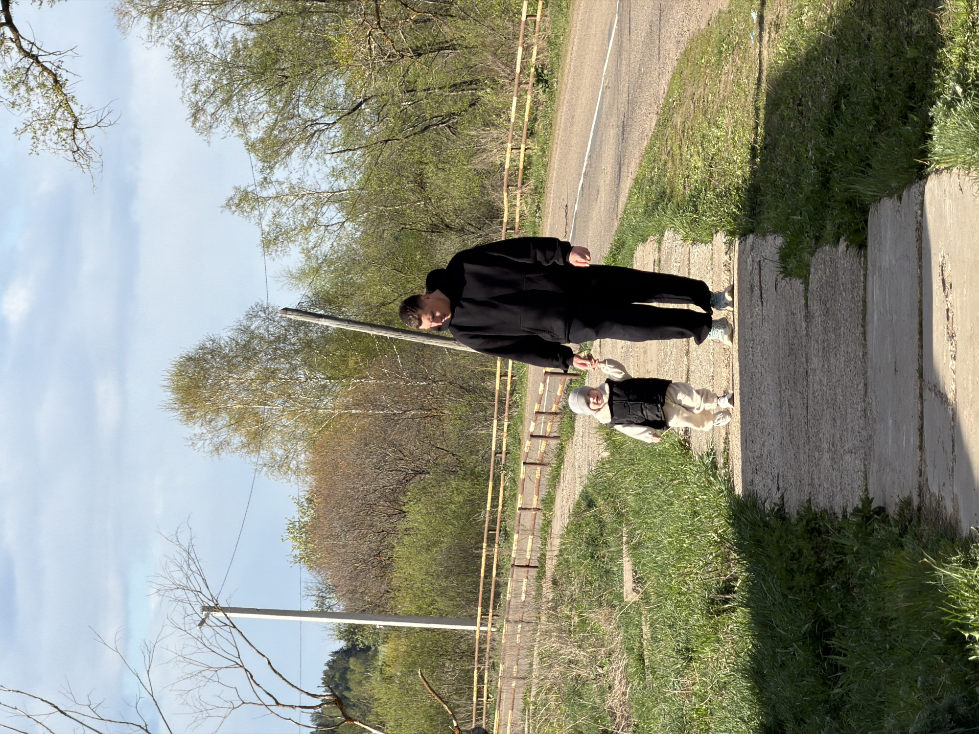 A medium shot, taken outdoors in daylight, shows a male adult holding the hand of a child, presumably walking. They are on a concrete path adjacent to a grassy area and a road. The adult male is dressed in a black hooded sweatshirt and black pants. The child is wearing lighter-colored clothing and a vest. The location is most likely in Большой Букор, Russia. Background scenery consists of trees, a fence and power lines.