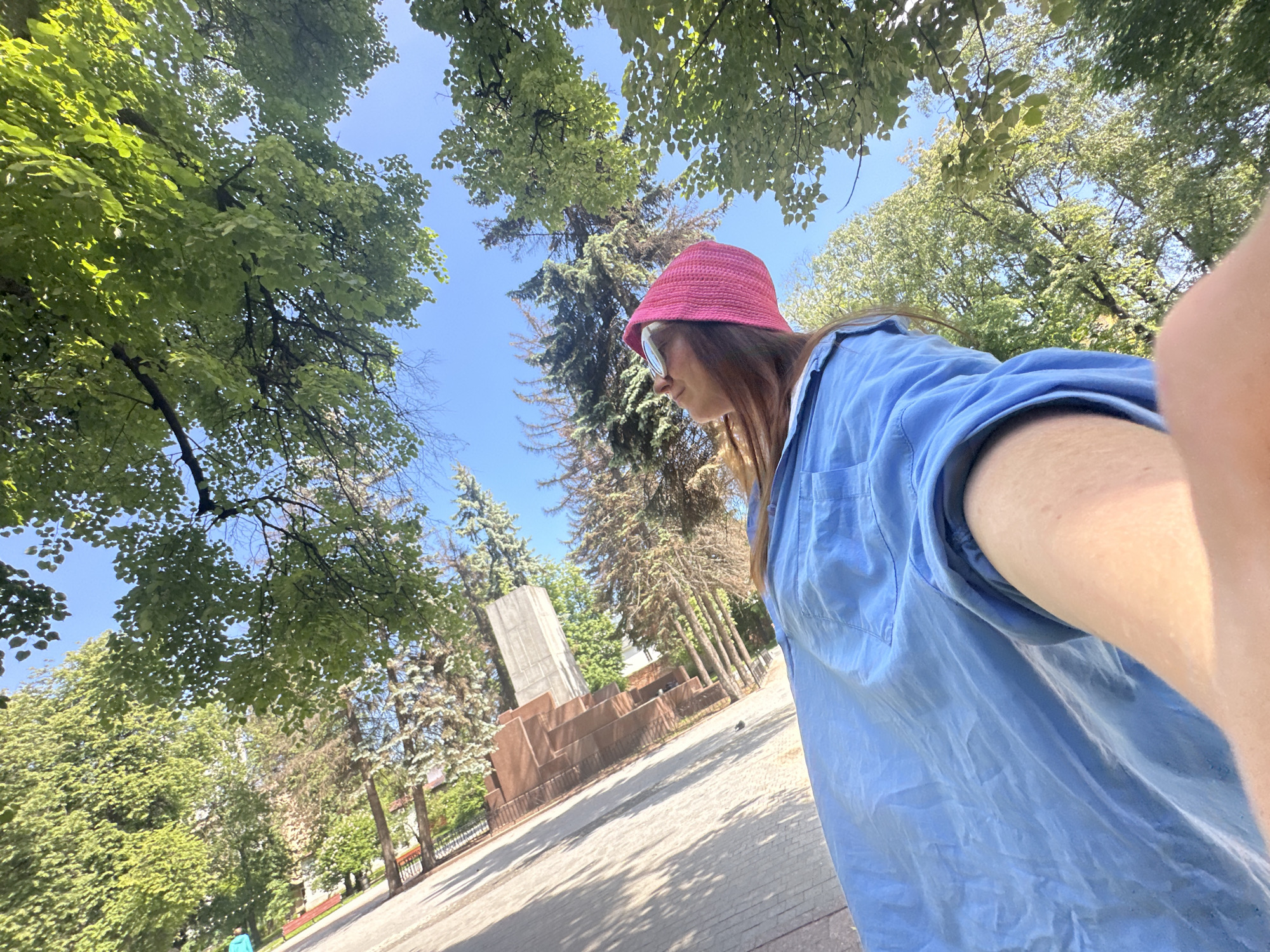 A low-angle selfie captures a person in an outdoor public space, likely a park or square, under a bright blue sky. The camera looks upwards, showcasing lush green tree canopies in the foreground and background, with sunlight dappling through the leaves. The person, positioned on the right side of the frame, wears a bright pink crocheted bucket hat, round sunglasses, and a light blue oversized long-sleeved shirt. Their arm and shoulder are visible on the far right, suggesting they are holding the camera.
In the mid-ground, a substantial stone monument is visible. It features a broad base of stepped, reddish-brown stone blocks, topped by a taller, lighter grey rectangular block. Surrounding the monument are various conifer trees, possibly spruces or firs, interspersed with deciduous trees. The ground is paved, and shadows from trees indicate strong overhead sunlight, suggesting it is midday or early afternoon on a clear, sunny day. A red park bench and a fence are faintly visible in the distance, further confirming a park setting. Based on the provided context, the scene is located in Nizhny Novgorod, Russia, and the monument appears to be a local landmark. No text is visible in the image.