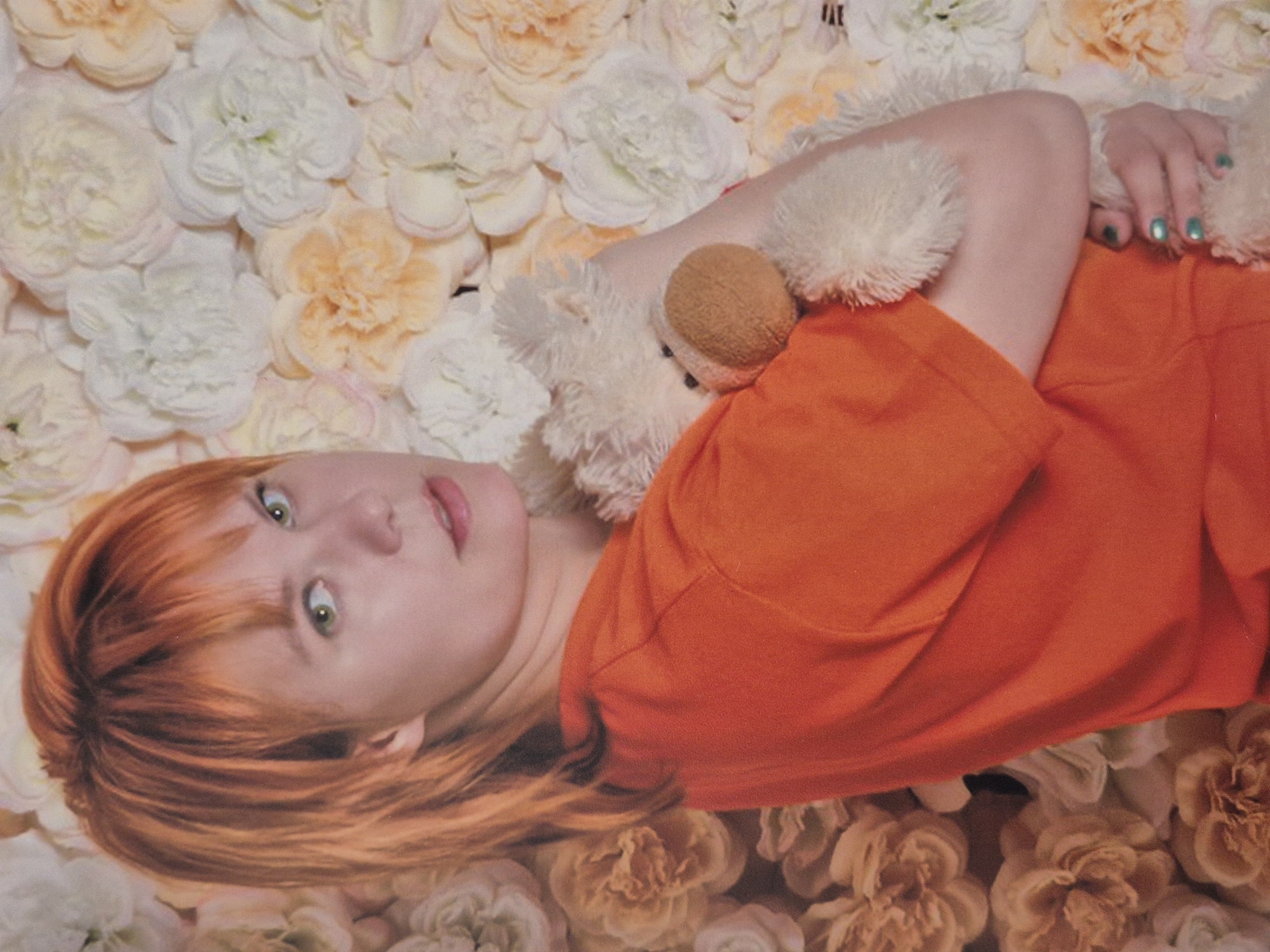 In the city of Schwäbisch Gmünd, Germany, an individual with short, orange hair, wearing an orange t-shirt, is holding a white teddy bear. The individual's face is partially visible, and their eyes are green. They are positioned in front of a backdrop of artificial flowers in shades of white and light orange. The teddy bear is fluffy and appears to be cradled by the individual. The lighting suggests an indoor setting, possibly a studio or a decorated room. The composition is a medium shot, focusing on the individual and the teddy bear.