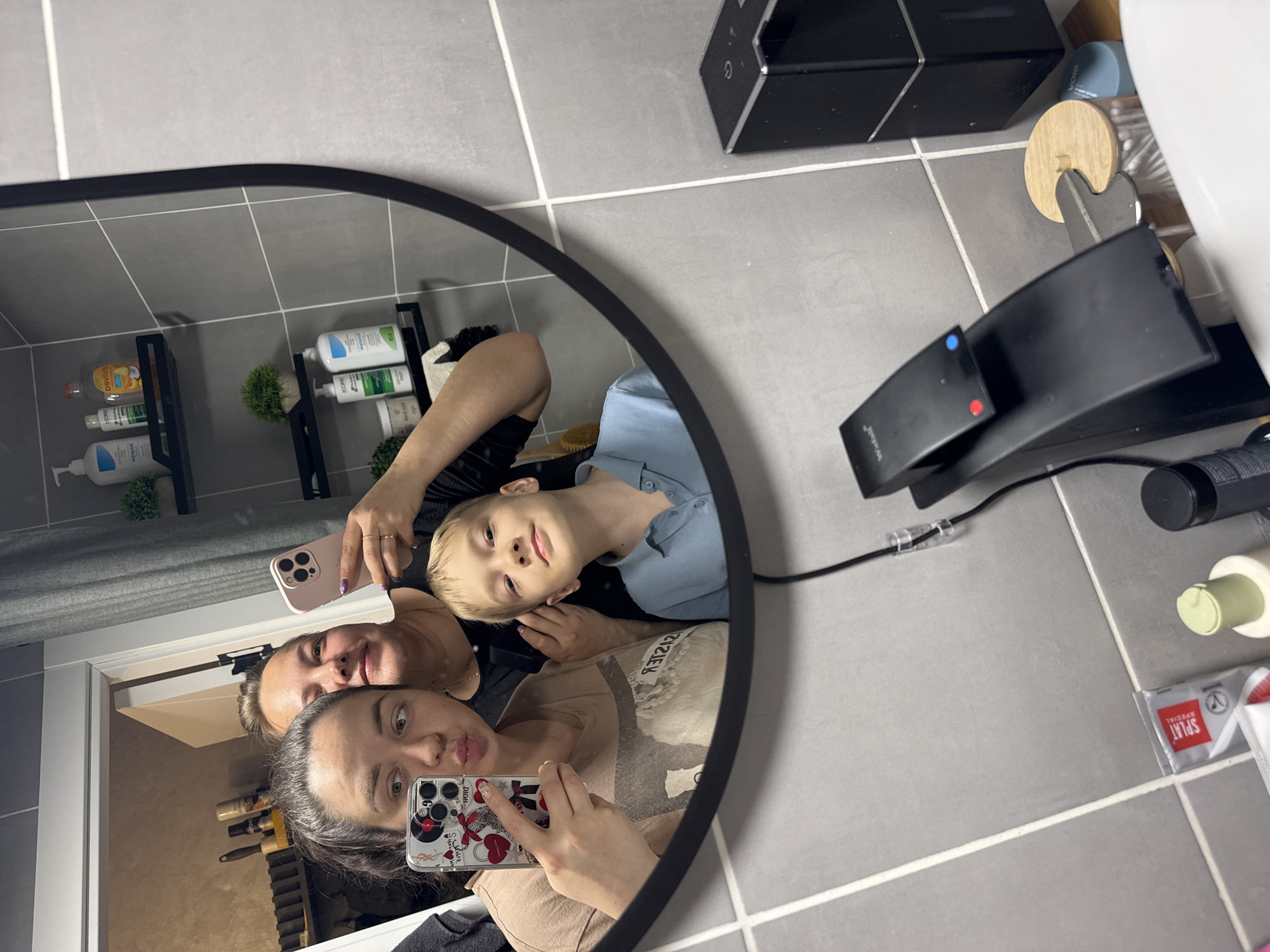 The image depicts a reflection in a round mirror of three people taking a selfie in what appears to be a bathroom. The foreground features a woman with dark hair, wearing a beige top, holding a phone with a Mickey Mouse case and pouting at the camera. Behind her is another woman holding a pink phone, and a young boy with blond hair is positioned between them. The bathroom has grey tiled walls and floor. A black shelving unit containing toiletries is visible in the background. Other objects in the bathroom include a black speaker, a toothbrush holder, a black hairdryer, and other toiletries on the floor. Based on the available data, it is not possible to verify the location as Yakhroma, Russia.