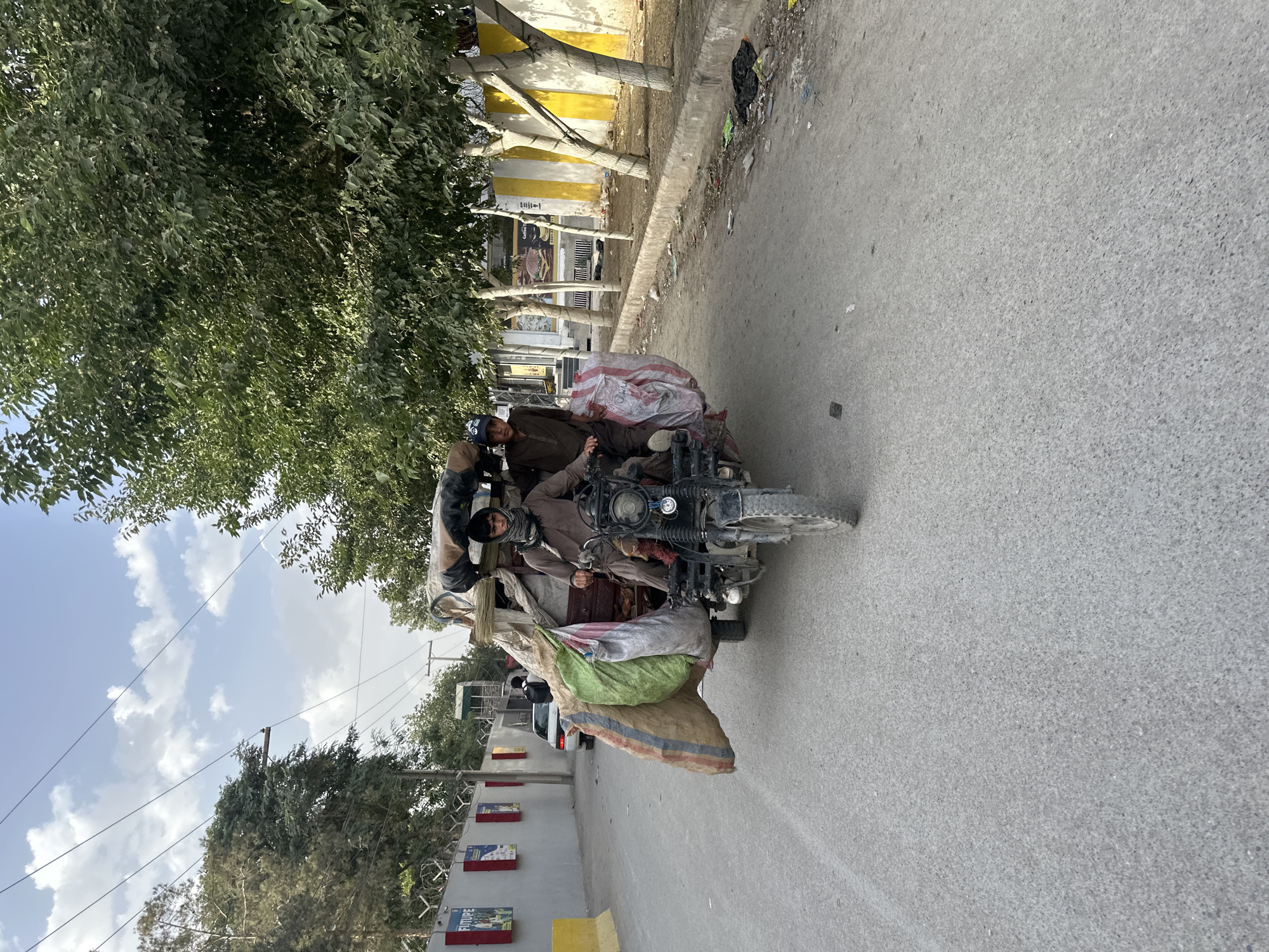 On an asphalt road in Quetta City Tehsil, Pakistan, a motorcycle-rickshaw hybrid transports multiple individuals and large sacks. The vehicle is positioned in the center of the road, appearing to be in motion. Two or more individuals are seated on the vehicle, with one person visibly driving. A variety of large sacks of materials are loaded on the vehicle. Buildings, foliage, and utility poles are present along the roadside. The weather appears clear and sunny.