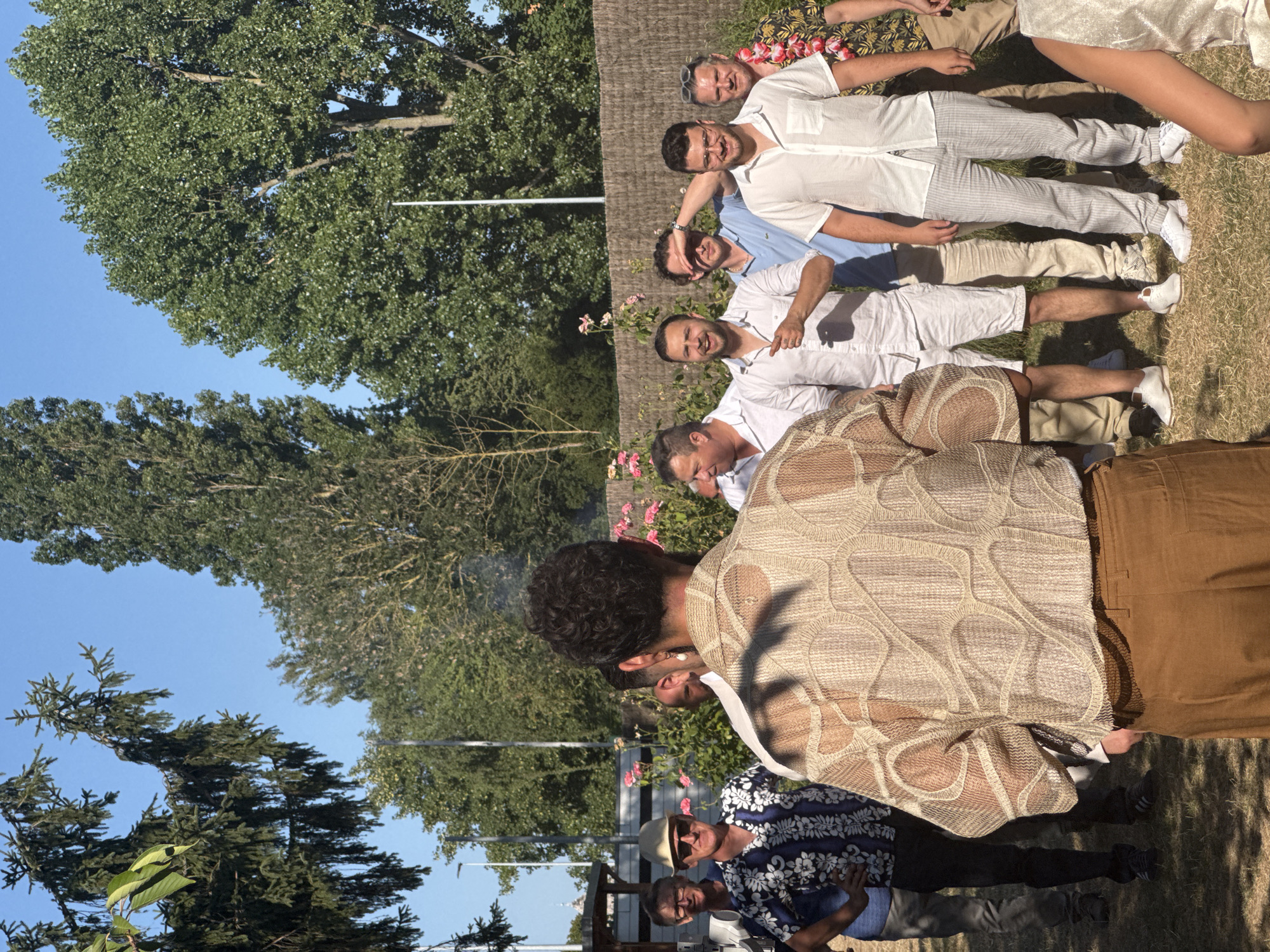 The image depicts a group of men gathered outdoors, with a clear blue sky and lush green trees in the background. The setting appears to be a backyard or park, given the presence of grass, a wooden fence, and rose bushes. The overall ambiance suggests a casual social gathering, possibly a barbecue or celebration.
In the foreground, a man is facing away from the camera, wearing a light-colored, textured, open-weave shirt and brown trousers. His presence dominates the immediate view, with his back turned to the viewer, obscuring some of the other individuals.
To his left, several men are visible. One man is wearing a blue and white patterned short-sleeved shirt, a straw hat, and sunglasses. Another man is in a white shirt and dark trousers. Further left, another man in a white shirt is partially visible.
To the right of the man in the foreground, a group of men are standing. One man in a white shirt and shorts is gesturing with his finger. Another man, wearing a light blue polo shirt, is shielding his eyes from the sun. Next to him, a man in a white short-sleeved shirt and light gray pinstriped trousers is smiling and holding a drink. To his right, a man with a colorful floral shirt and a lei around his neck is also visible.
The image was taken in Hem, France, and the specific location within Hem appears to be a private garden or outdoor event space. The lighting suggests it is daytime, likely late afternoon due to the warm, soft light and the long shadows cast on the grass. The men are dressed in a variety of casual attire, ranging from floral shirts to plain white shirts, with some wearing hats and sunglasses. The mood seems relaxed and convivial, with several individuals smiling and interacting. There are some subtle details such as the presence of a camera on a tripod on the left side of the frame, suggesting the event may have been professionally documented.